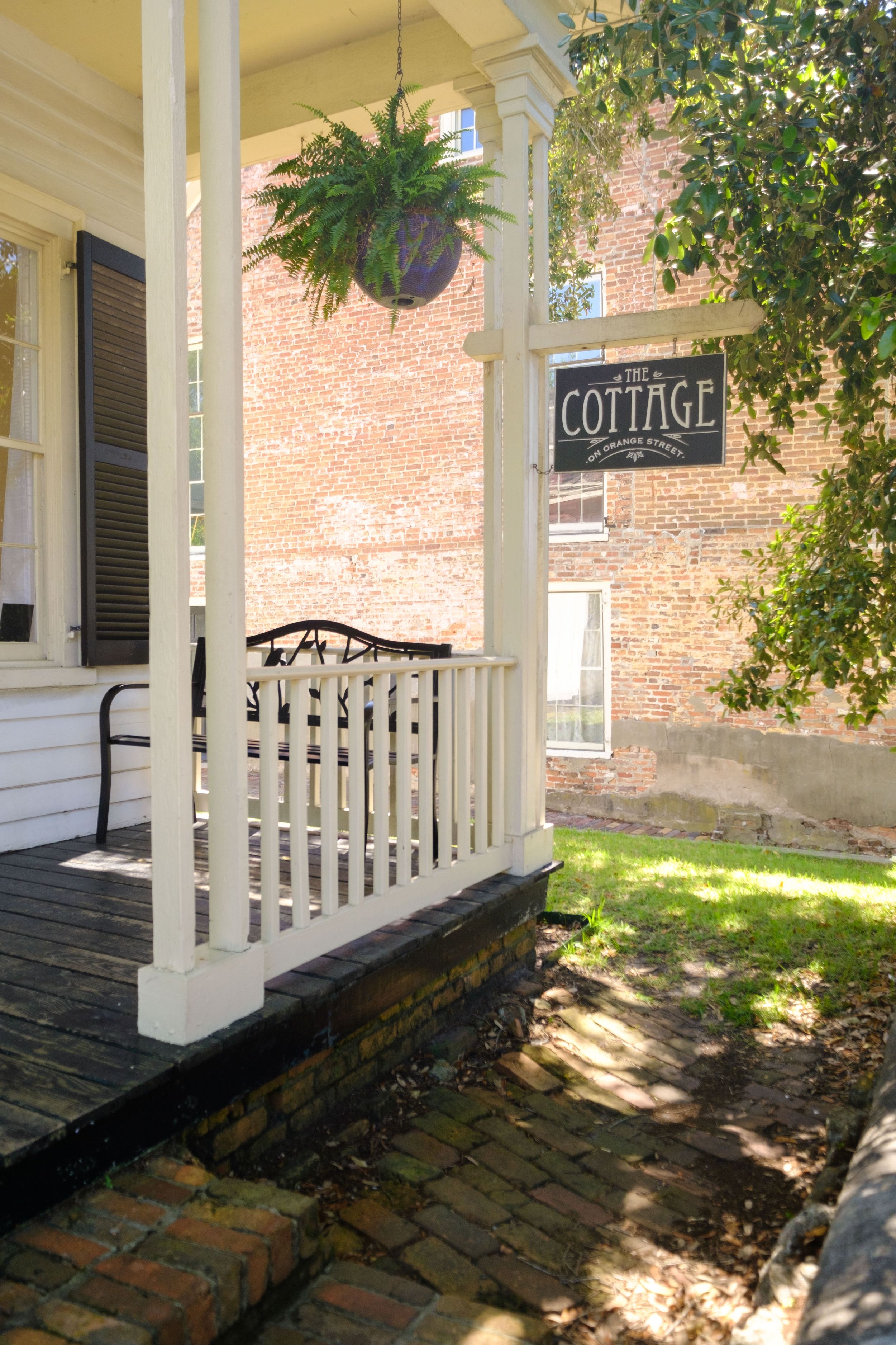 Close-up view of a wooden cottage entrance sign with decorative black text and trim against a sunlit brick wall.