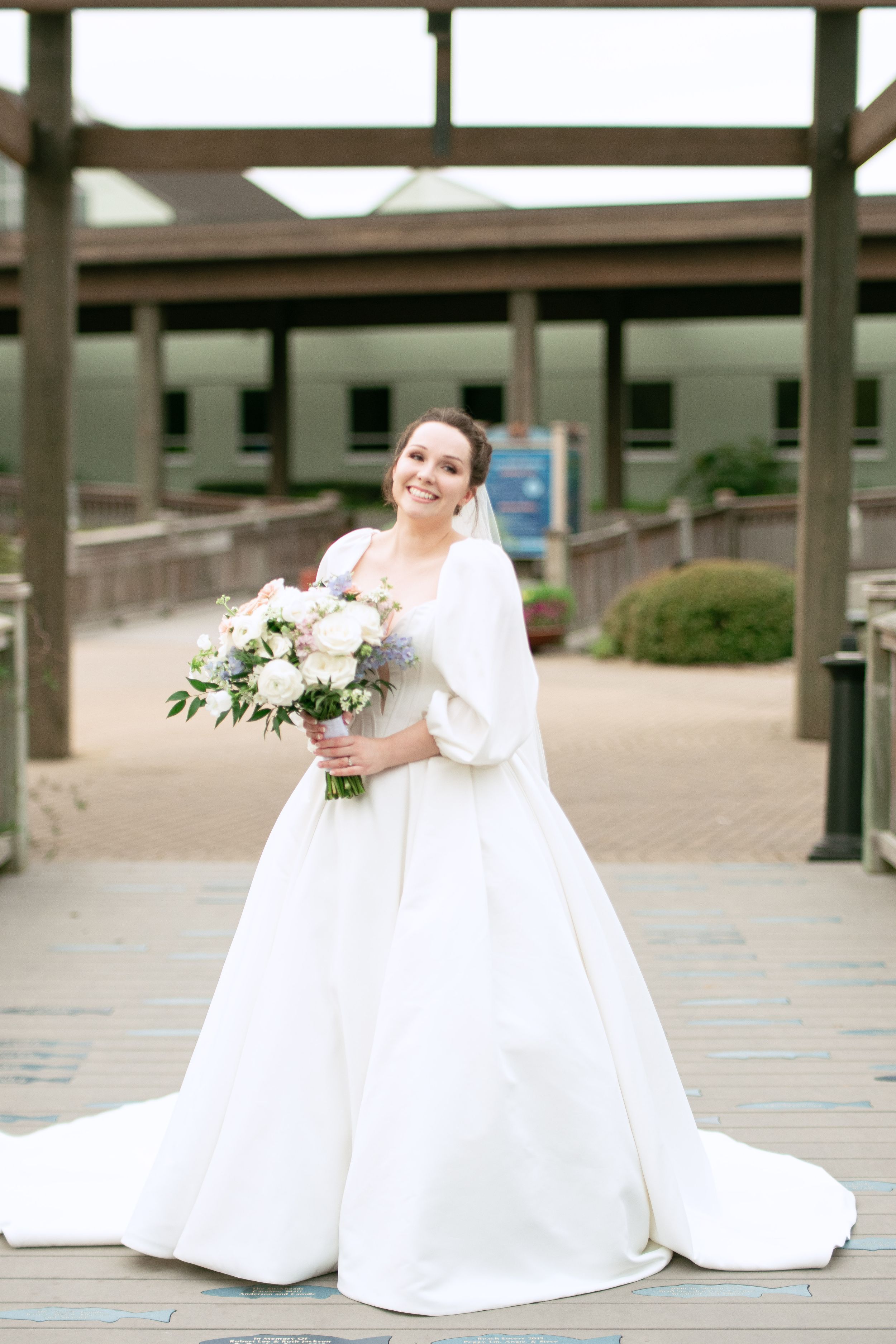 A bride in a classic white ballgown wedding dress stands on a wooden deck holding a lush white floral bouquet.