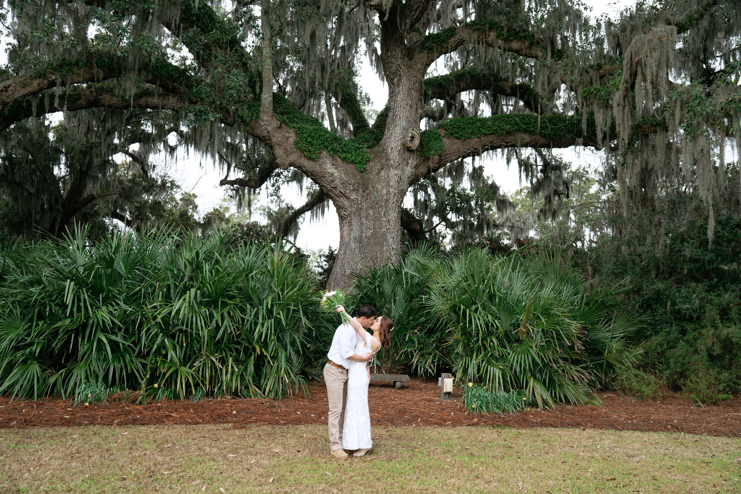 Two figures in white embrace beneath a majestic Spanish moss-draped oak tree in a lush garden setting.