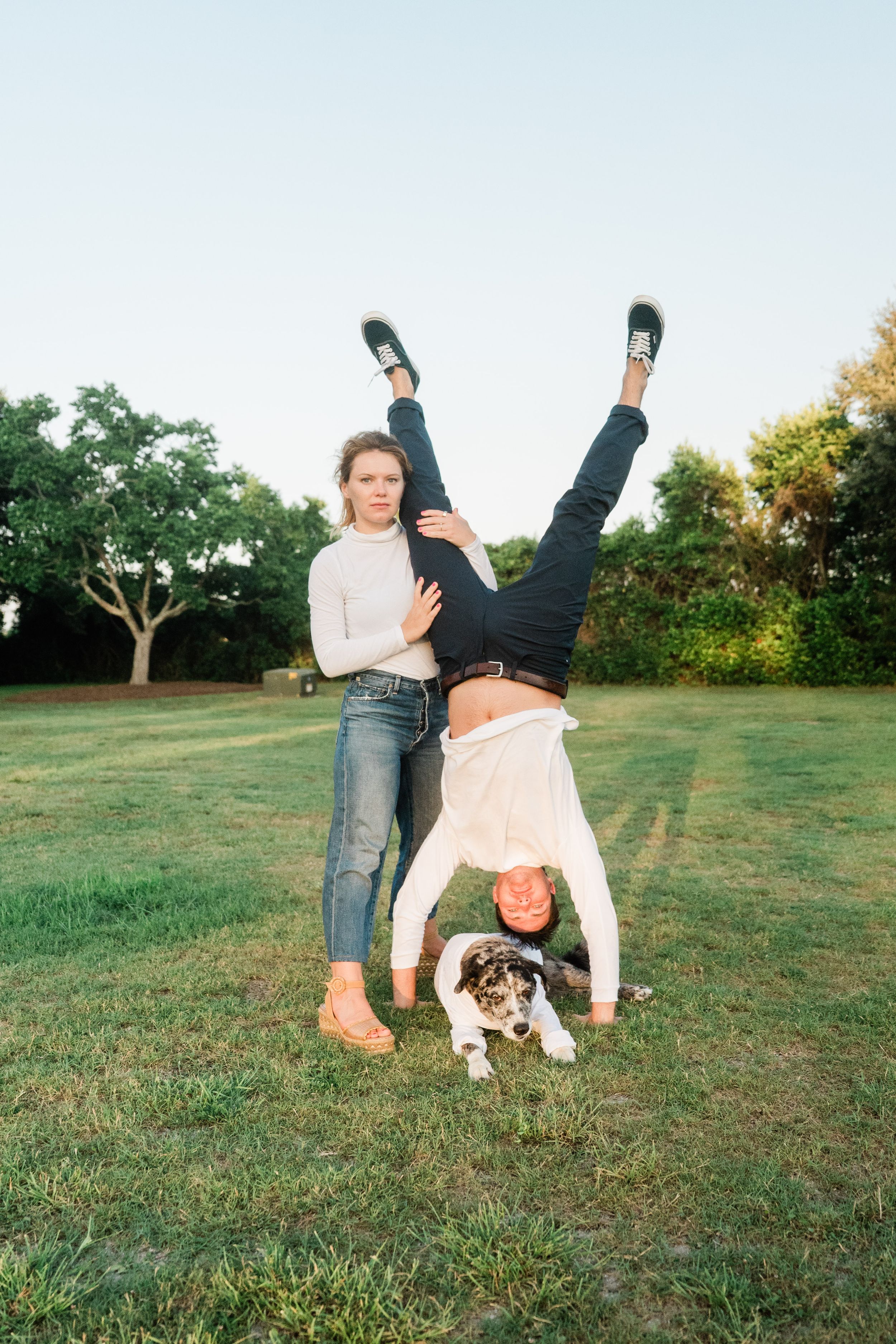 Playful scene on a grassy lawn with two people doing a handstand pose while a small dog sits beneath them at sunset.