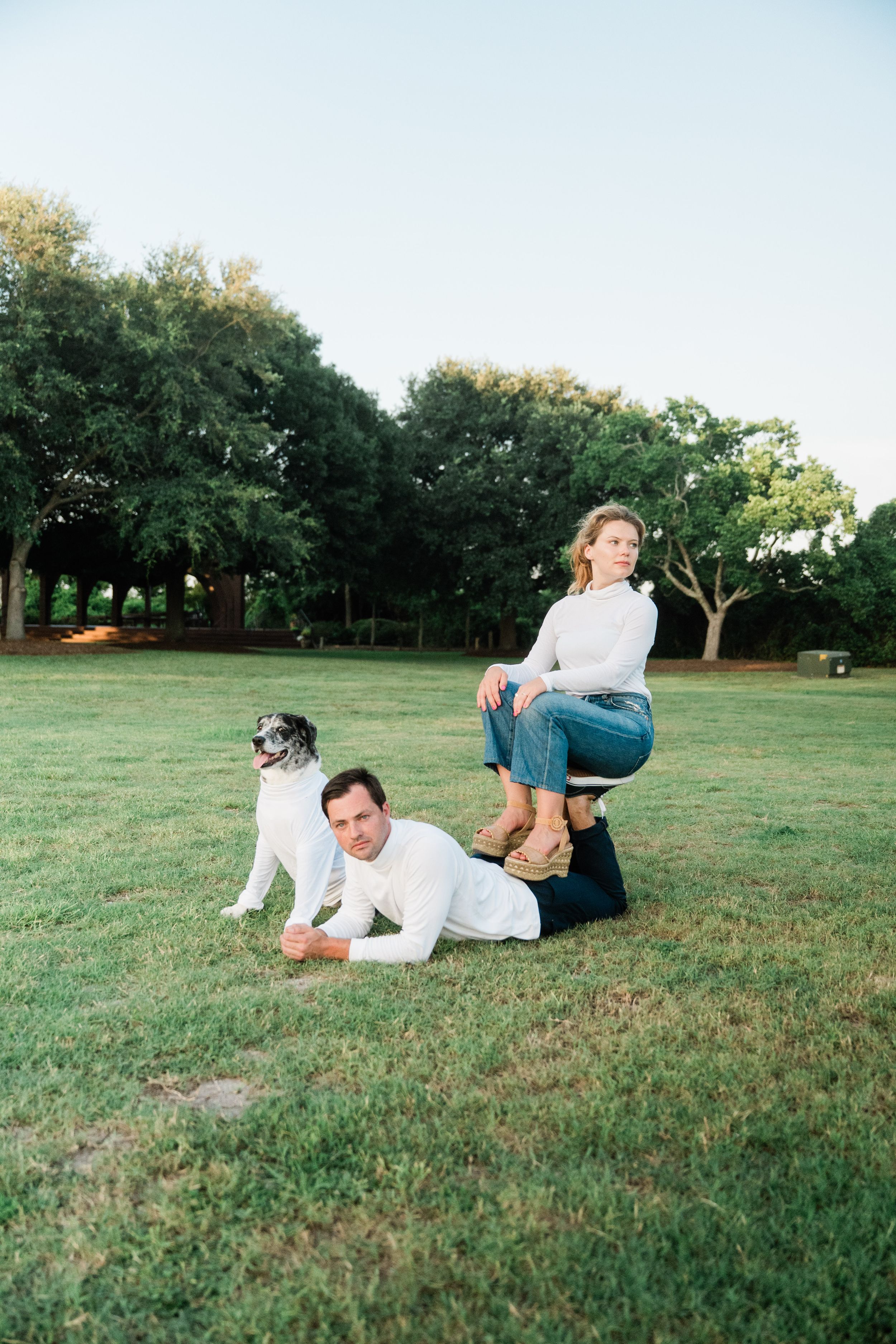 Two people and their dog create a fun acrobatic pose on a grassy lawn with trees in the background.