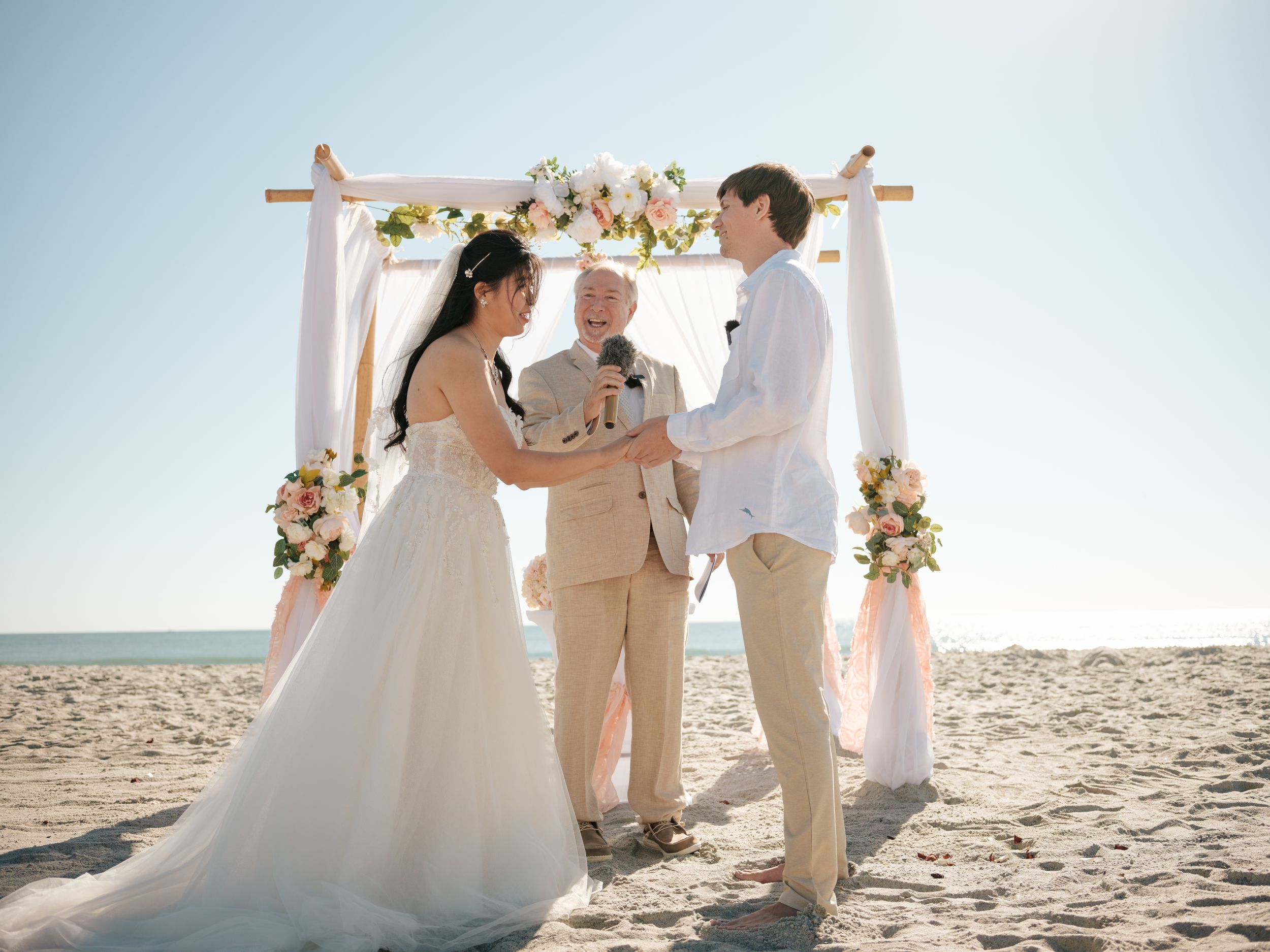 Beach wedding ceremony with floral arch decorations set against ocean backdrop in soft sunlight.