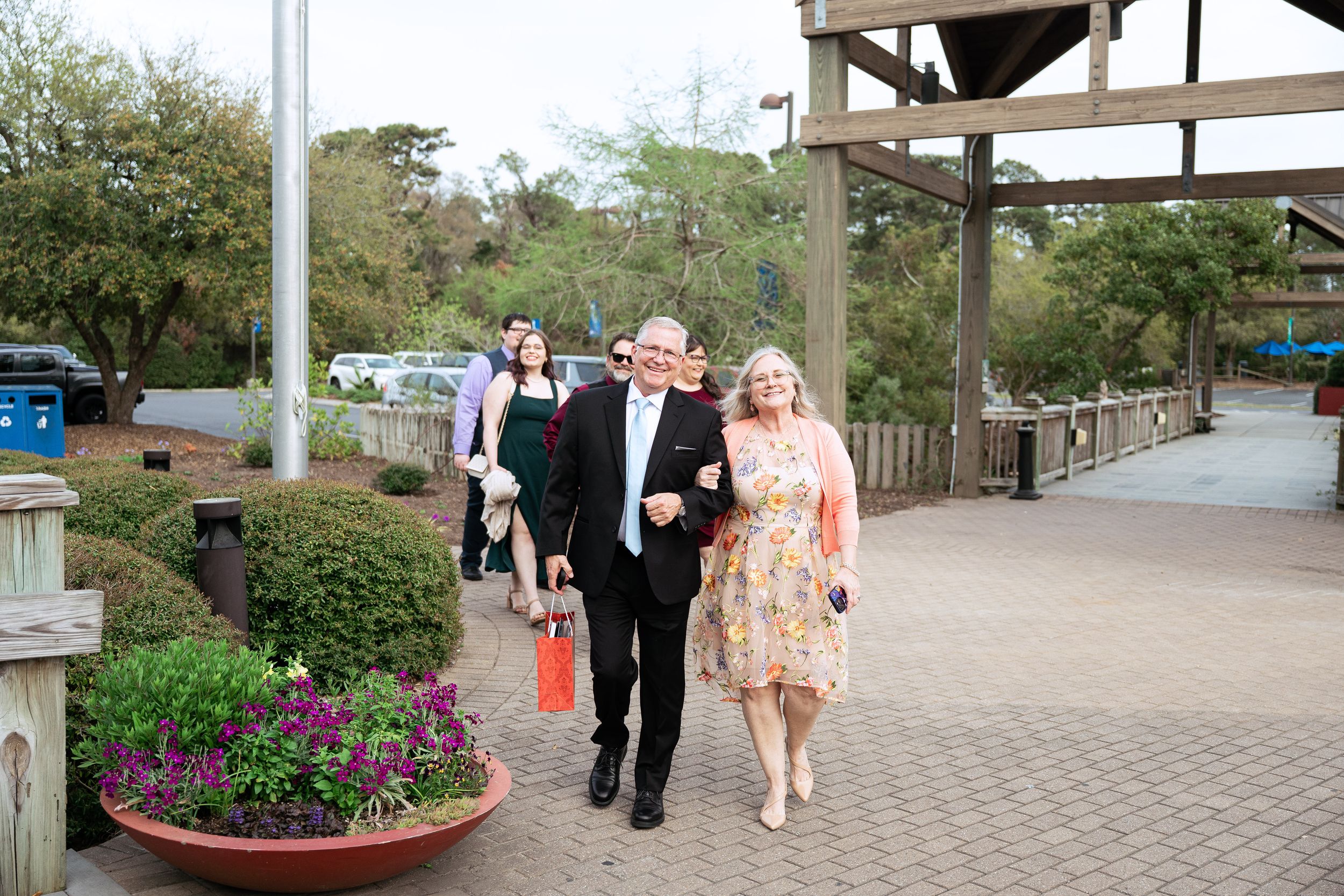 Wedding guests walk along a paved pathway decorated with colorful flower planters at an outdoor venue.