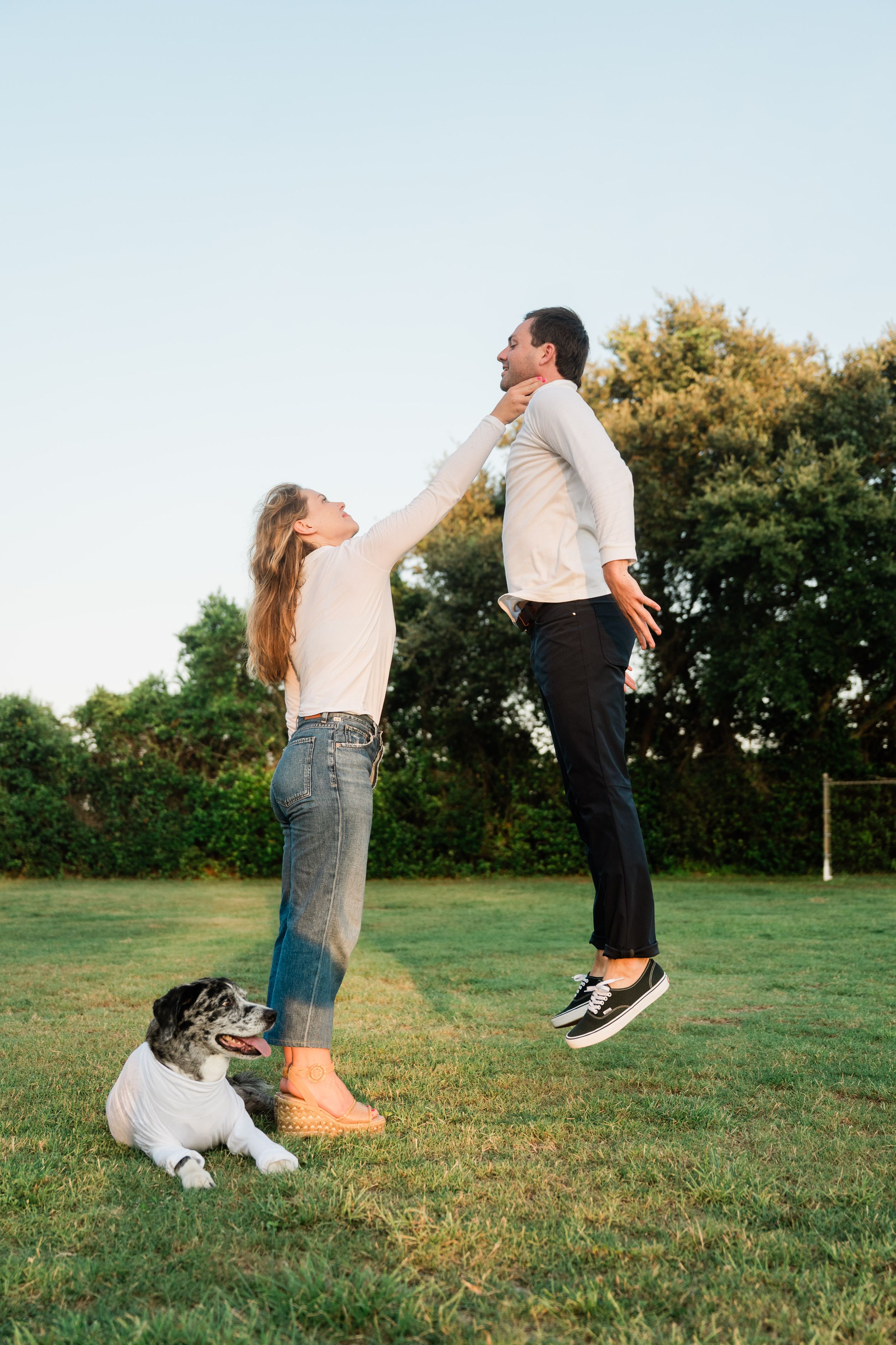 Energetic couple and their dog playing and jumping together on a grassy field during golden hour.