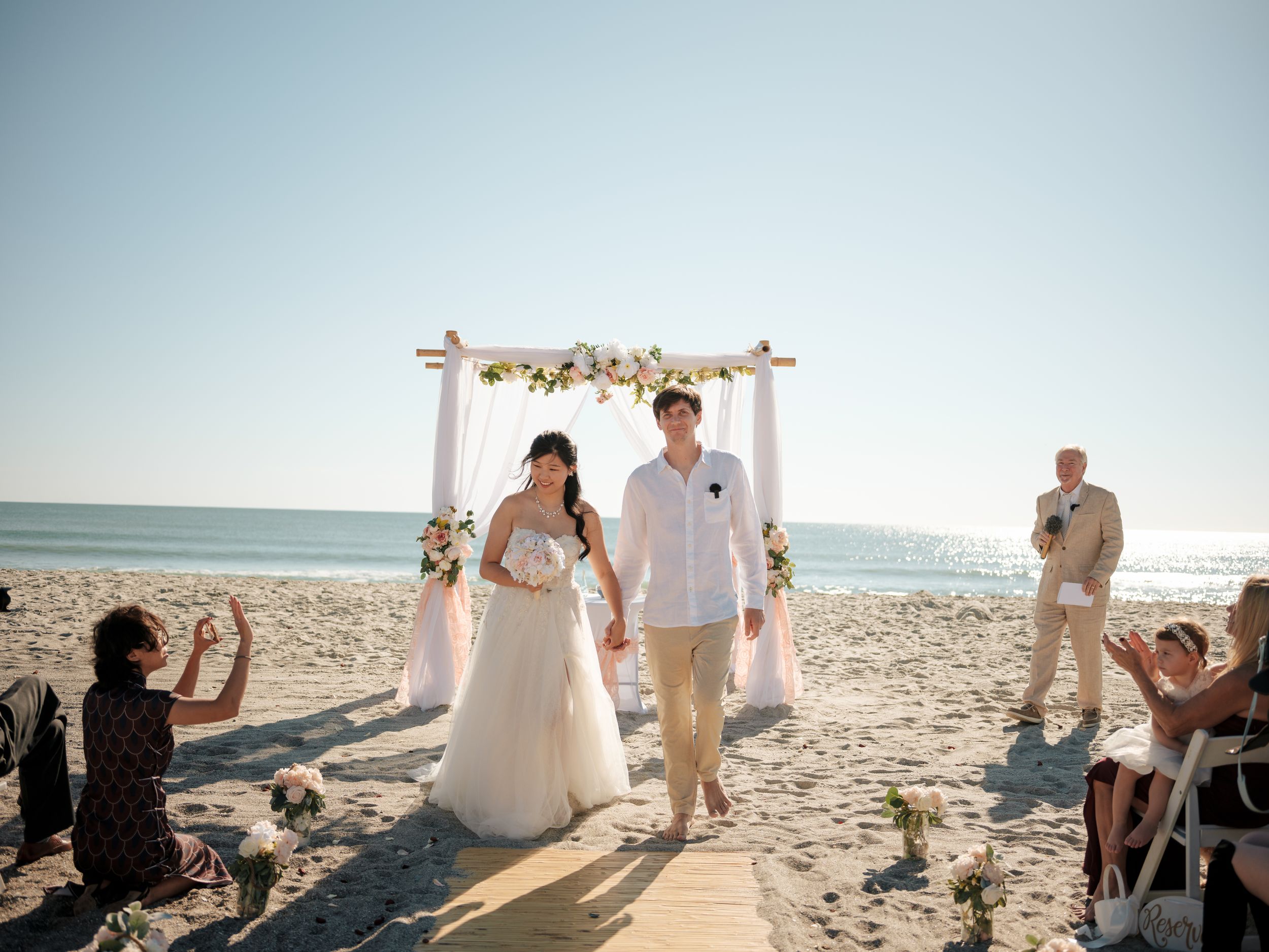 Wedding guests gather around a floral arch for a beach ceremony at sunset.