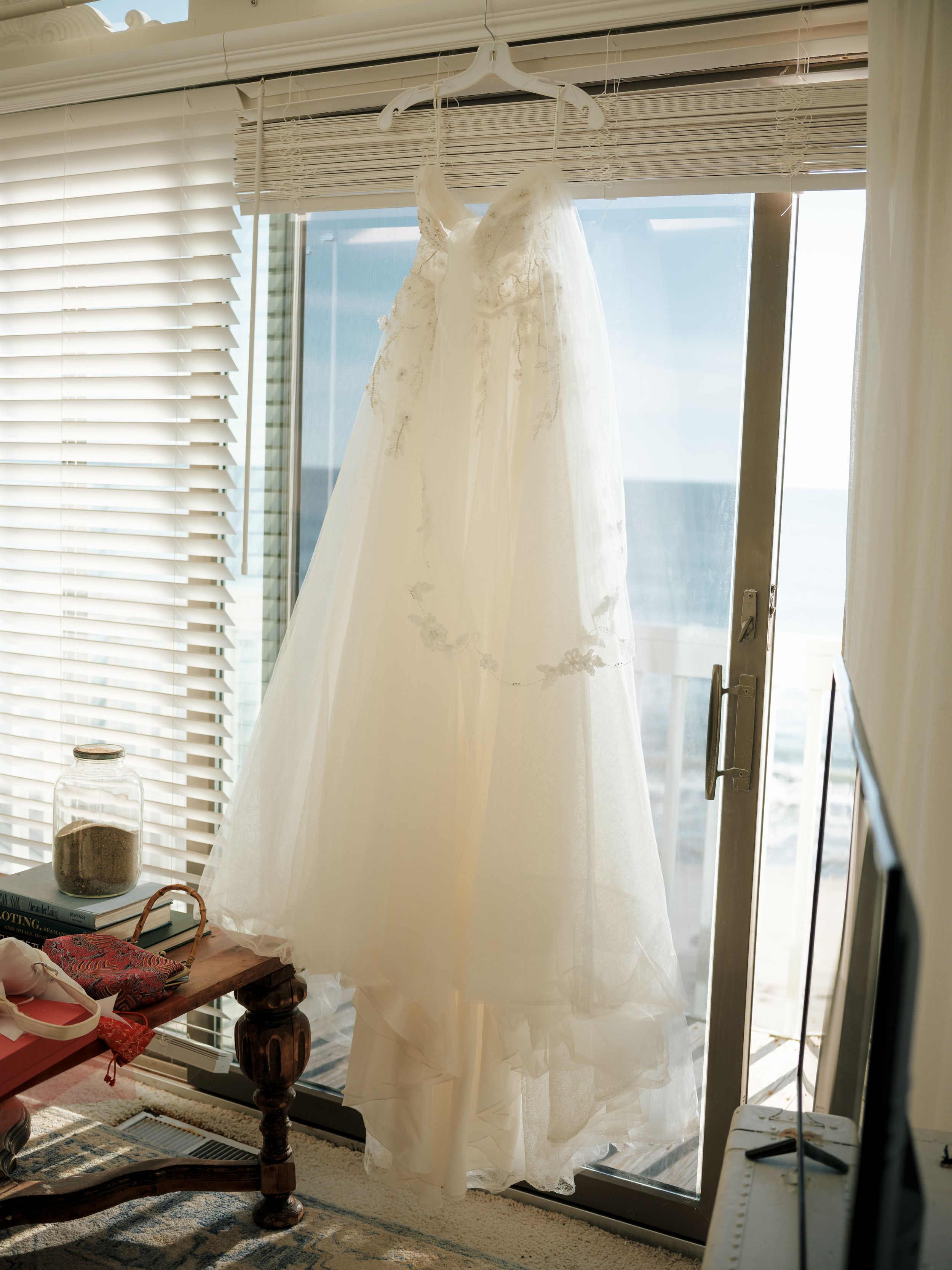 Several white wedding dresses hang near sunlit windows in a bright room.