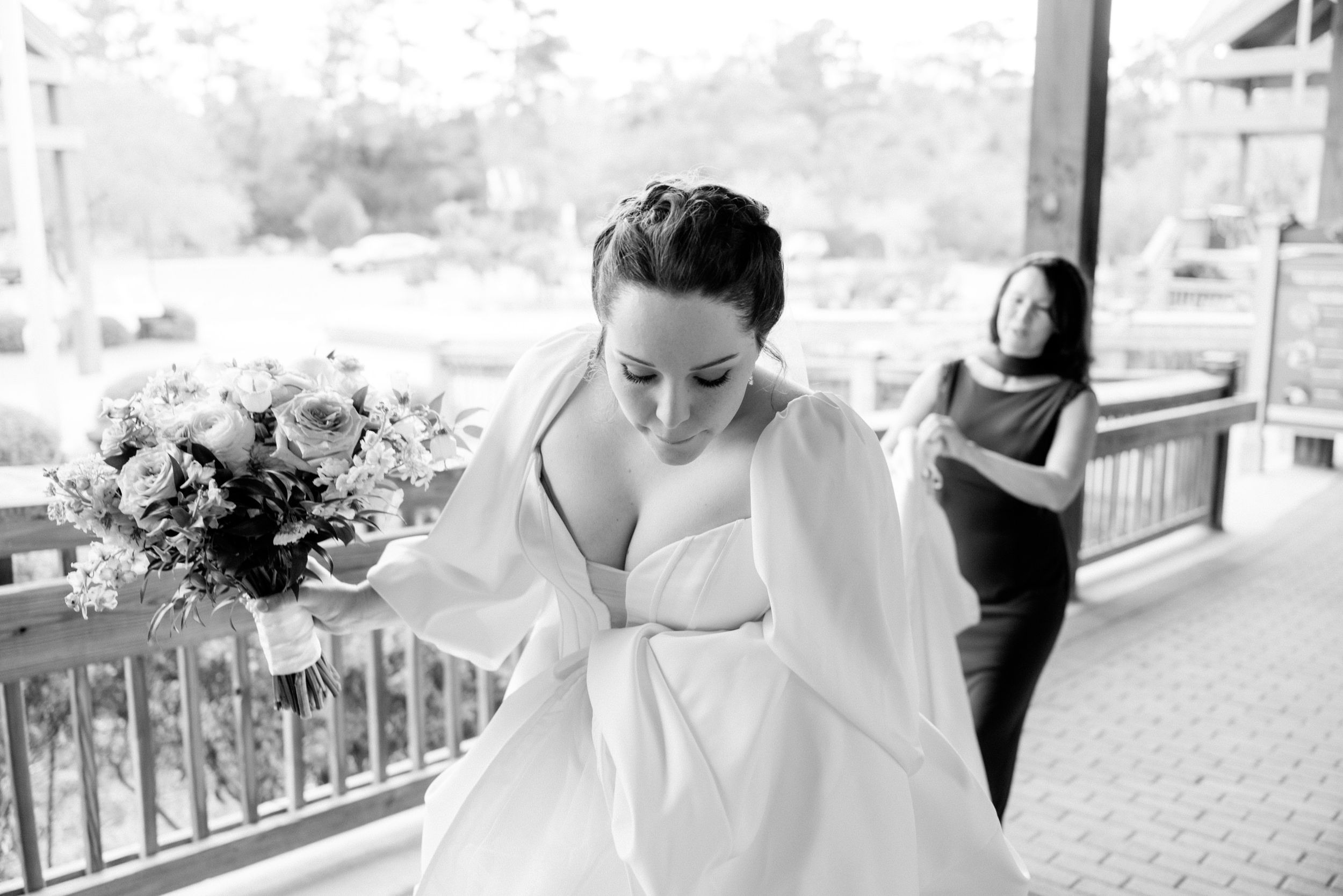 A flowing white wedding dress being adjusted on a covered porch with natural lighting.