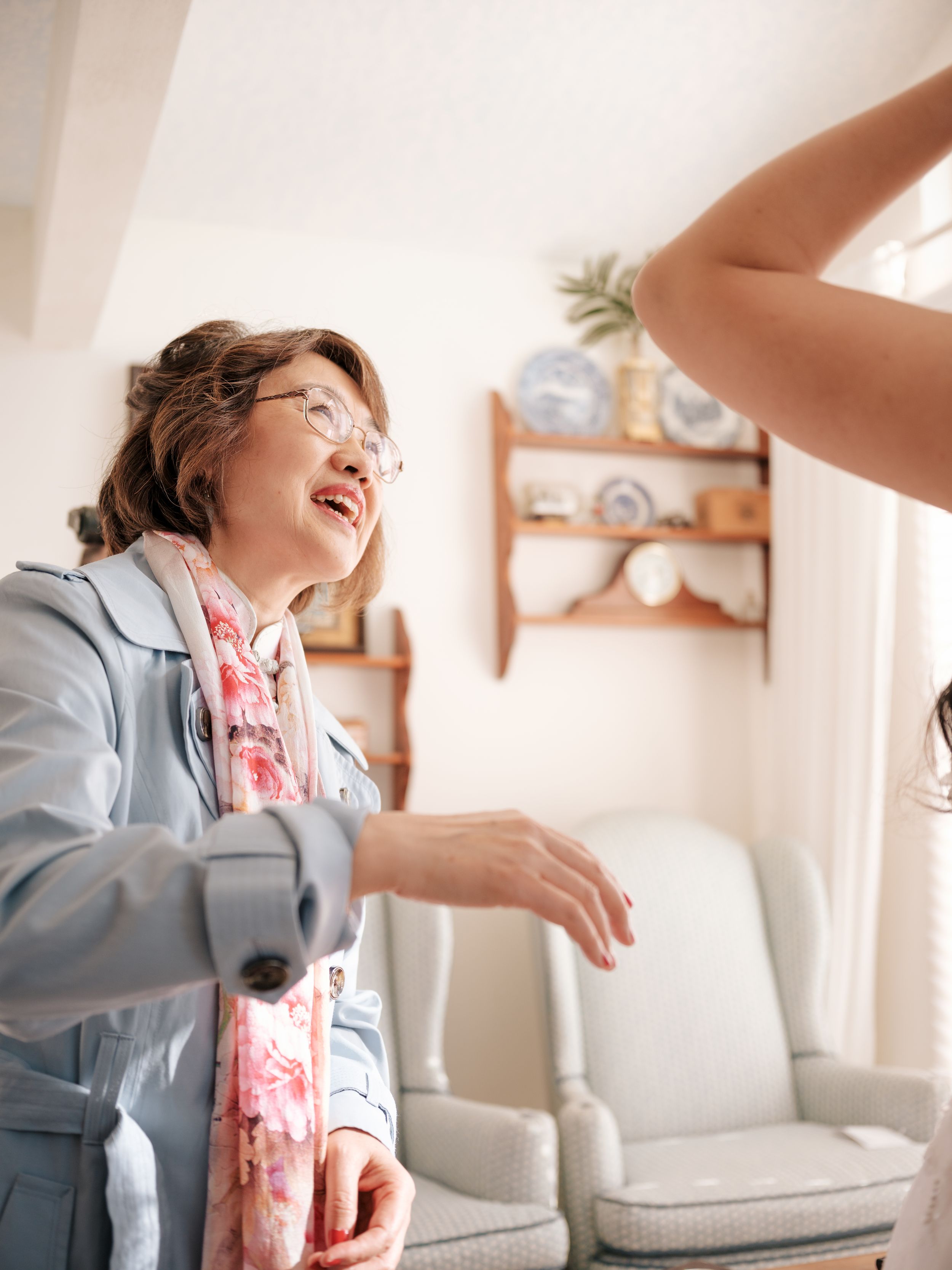 Woman in light blue jacket and floral dress dances joyfully in a bright indoor setting.