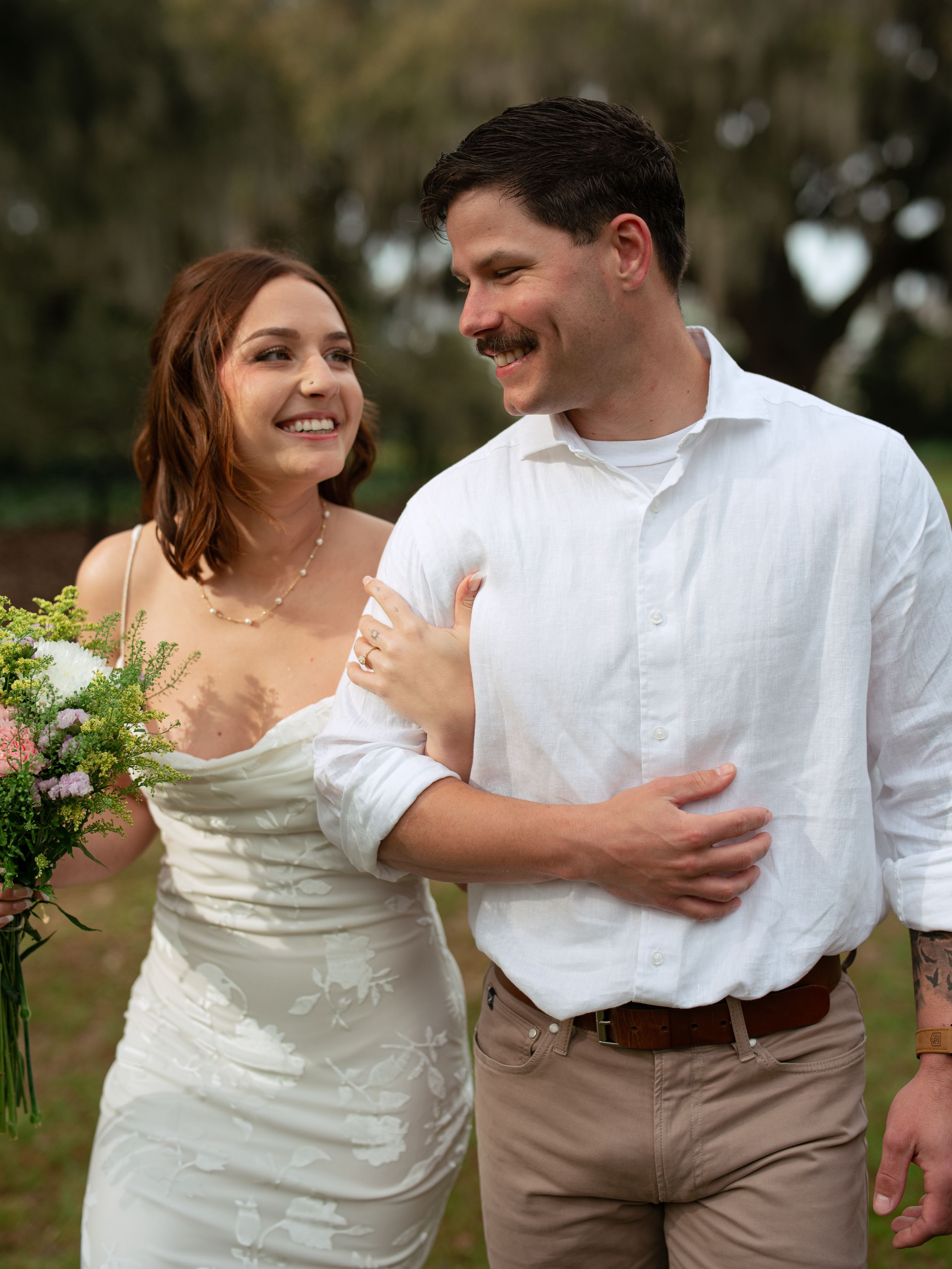 A joyful couple shares a candid moment outdoors while holding a bouquet of flowers.