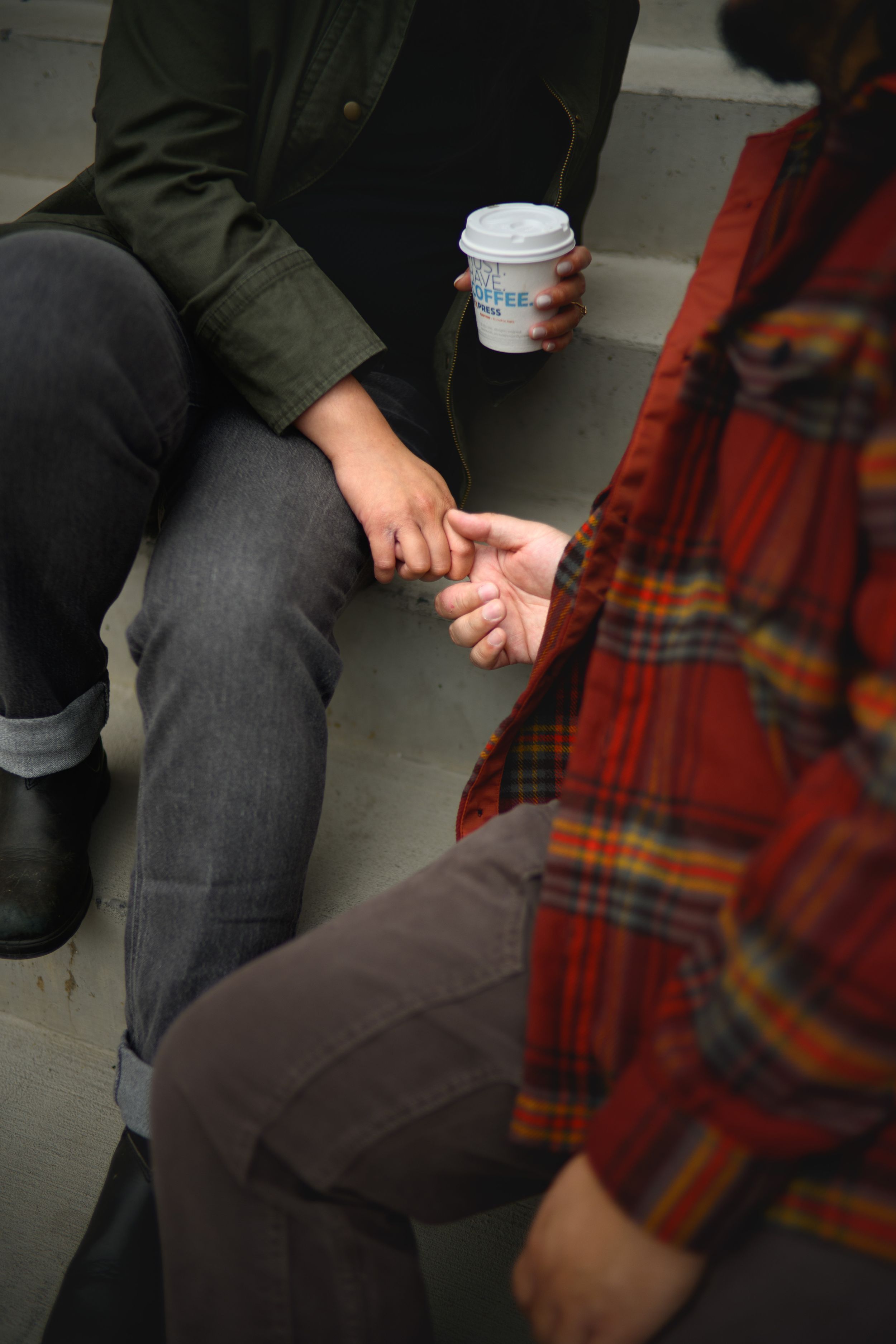 Two people sitting close together on a bench with one person holding a coffee cup in casual attire.