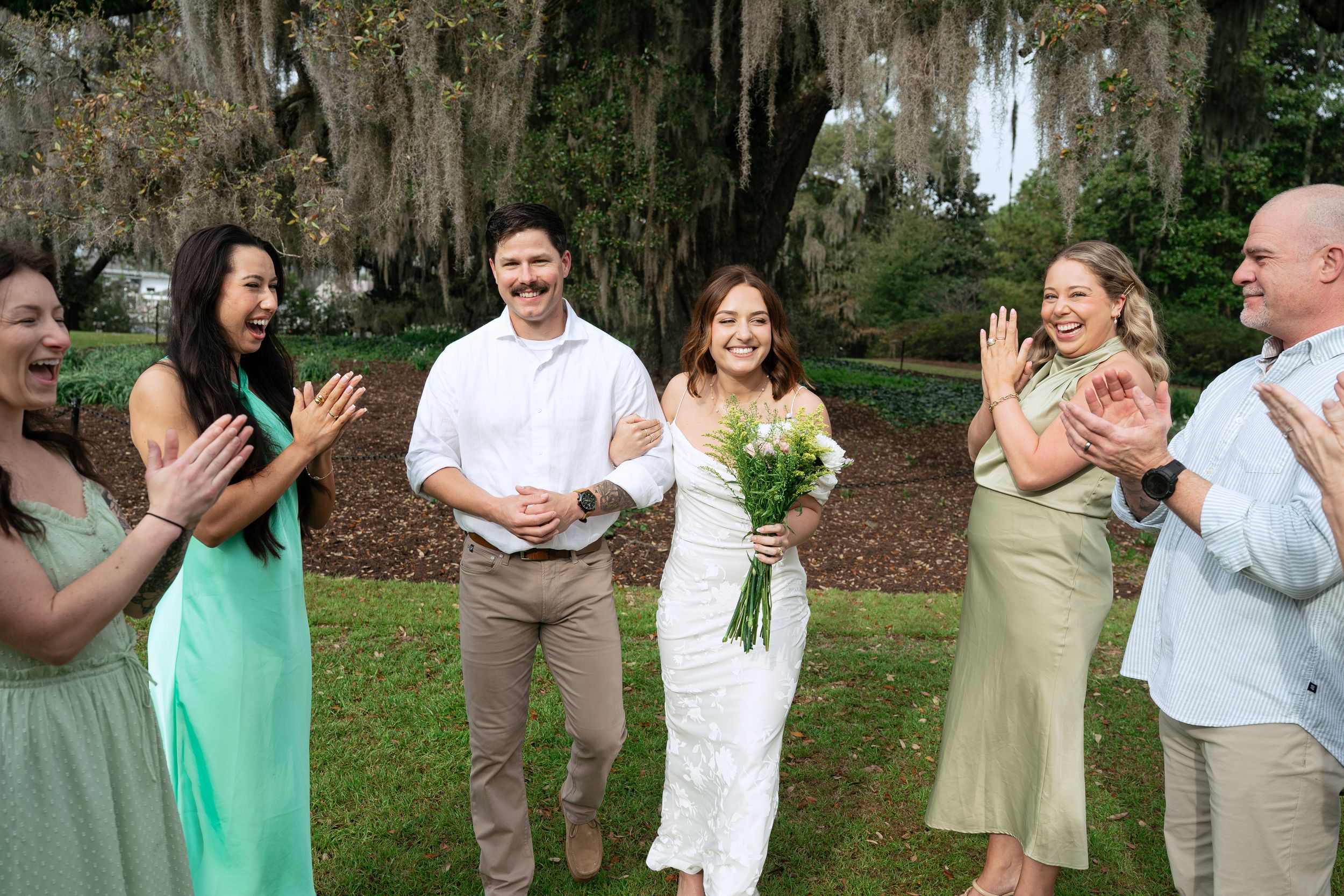 A group of wedding guests stands in a line outdoors laughing together while wearing green and white attire.