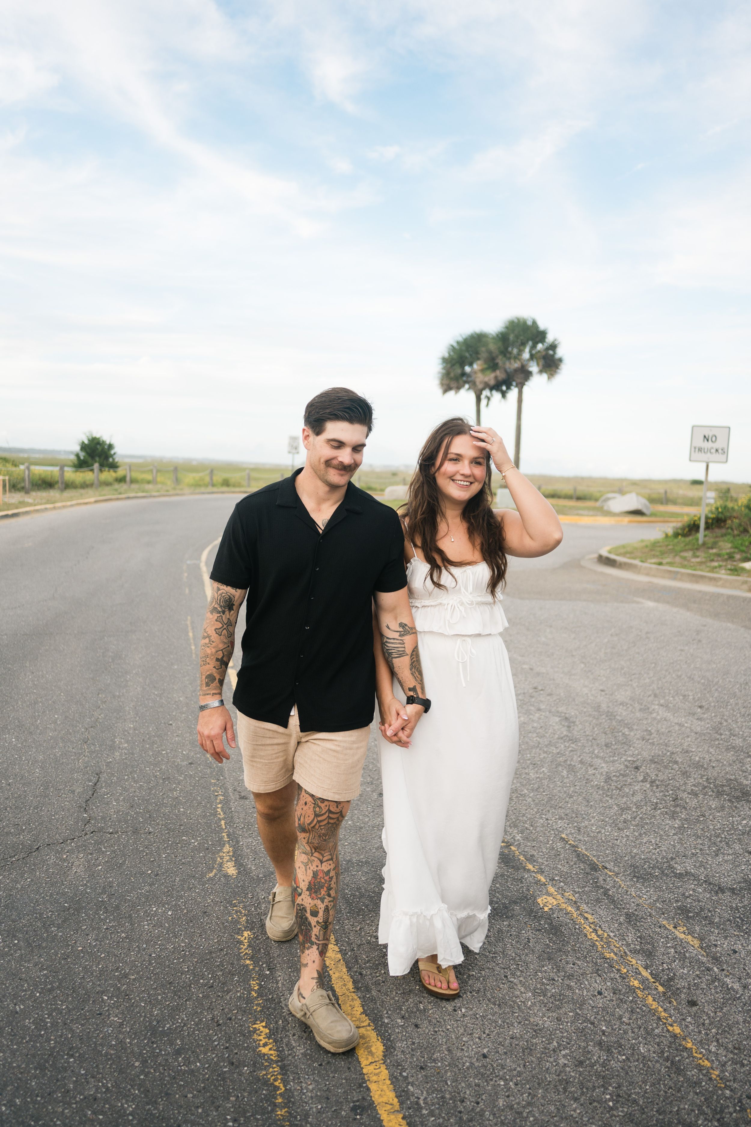 A couple walks hand in hand down a coastal road with palm trees and blue skies in the background.