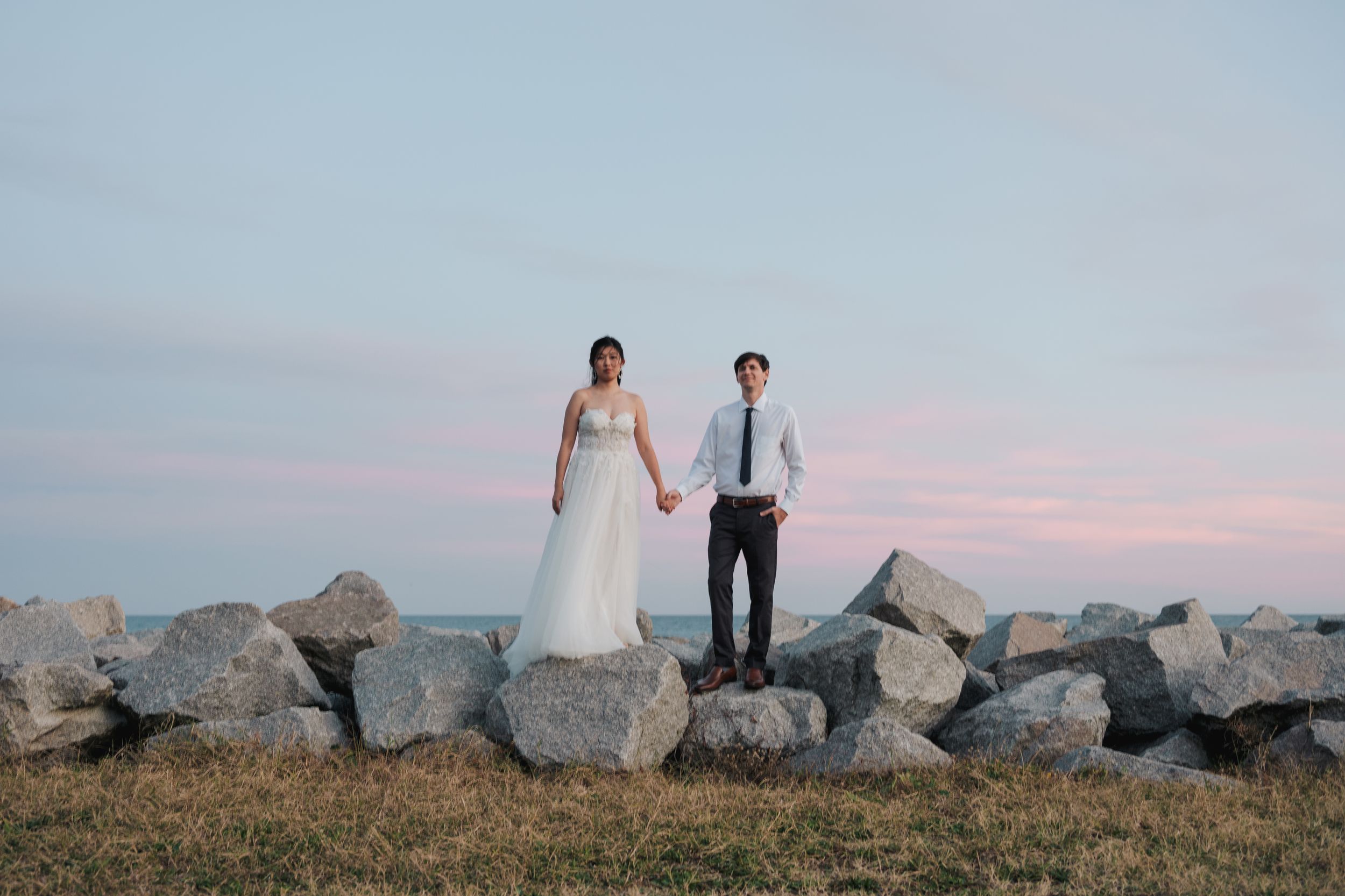 A couple in wedding attire walks along a rocky coastal shoreline during sunset creating a romantic silhouette.