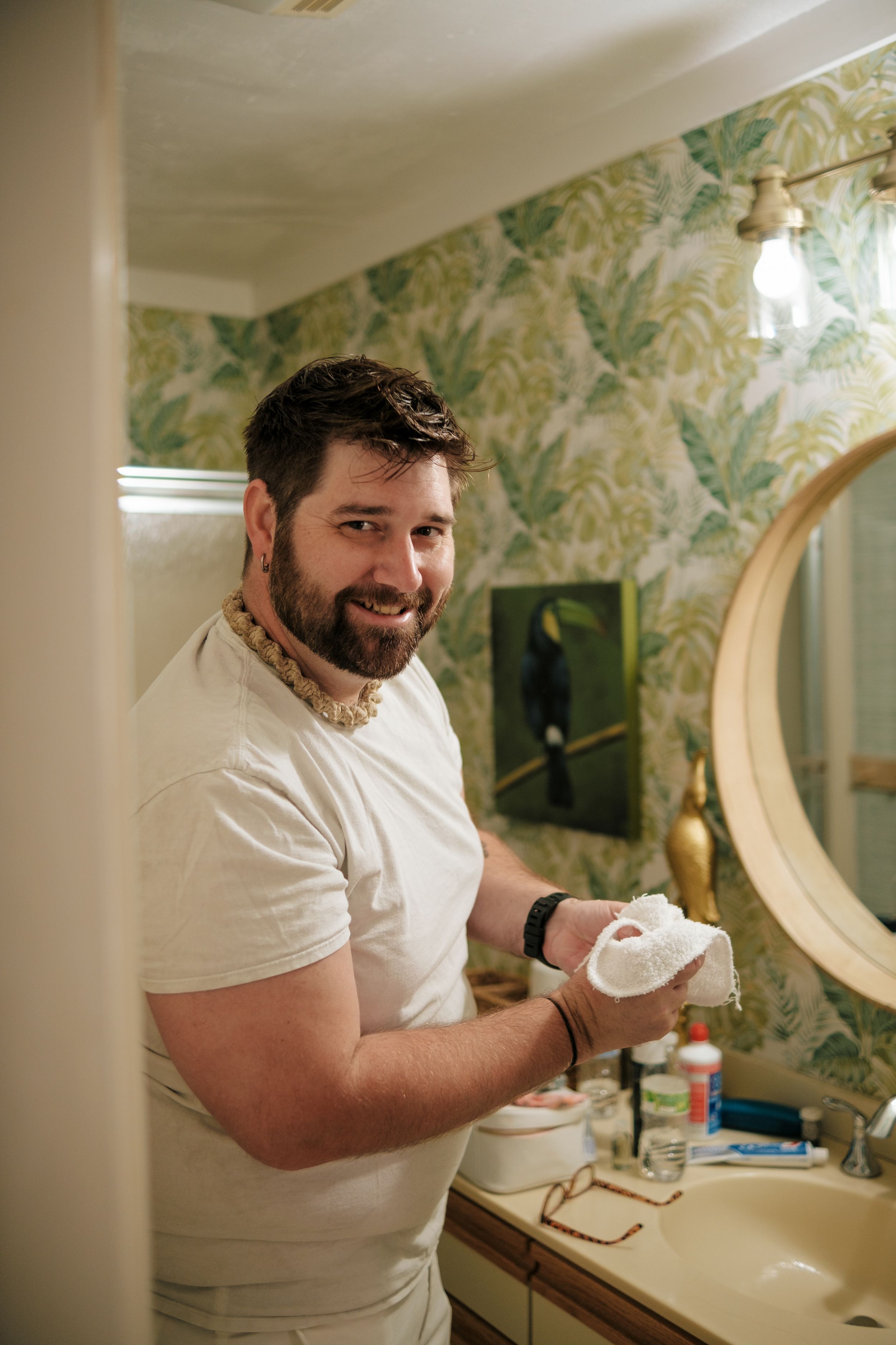 Person in white shirt smiling while getting ready in bathroom with floral wallpaper.