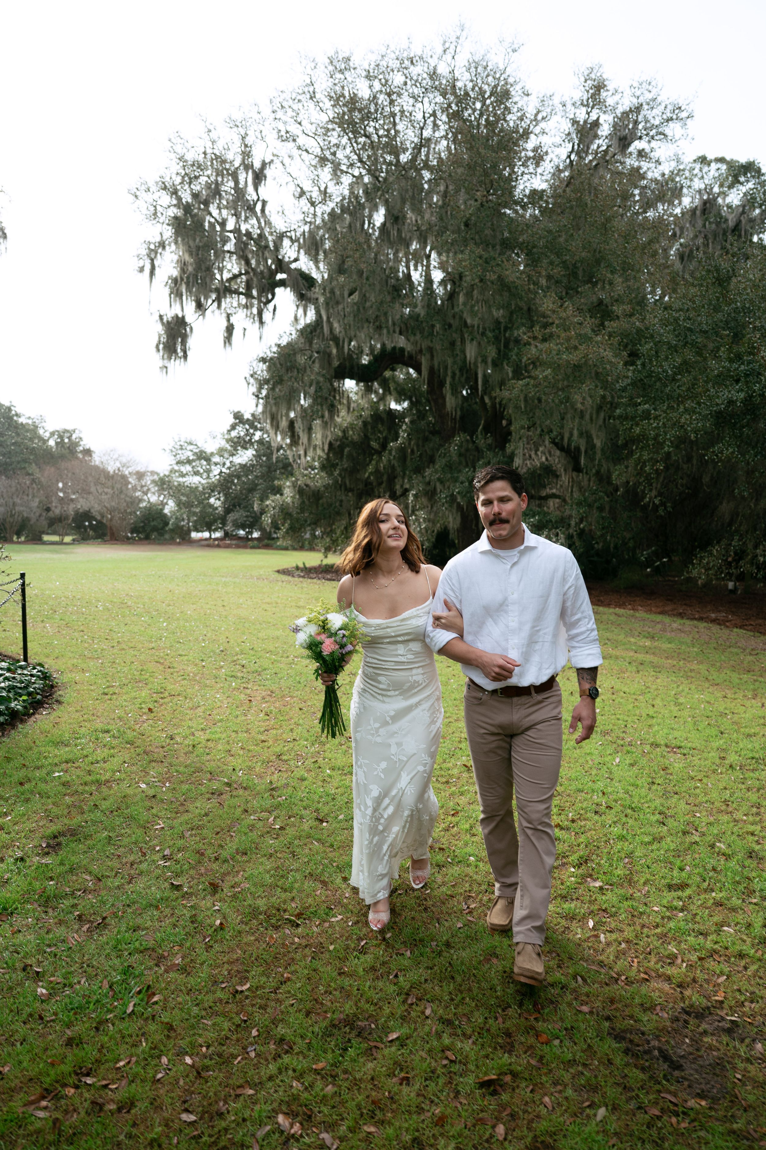 A couple walks hand in hand across a grassy lawn with Spanish moss draped trees in the background.