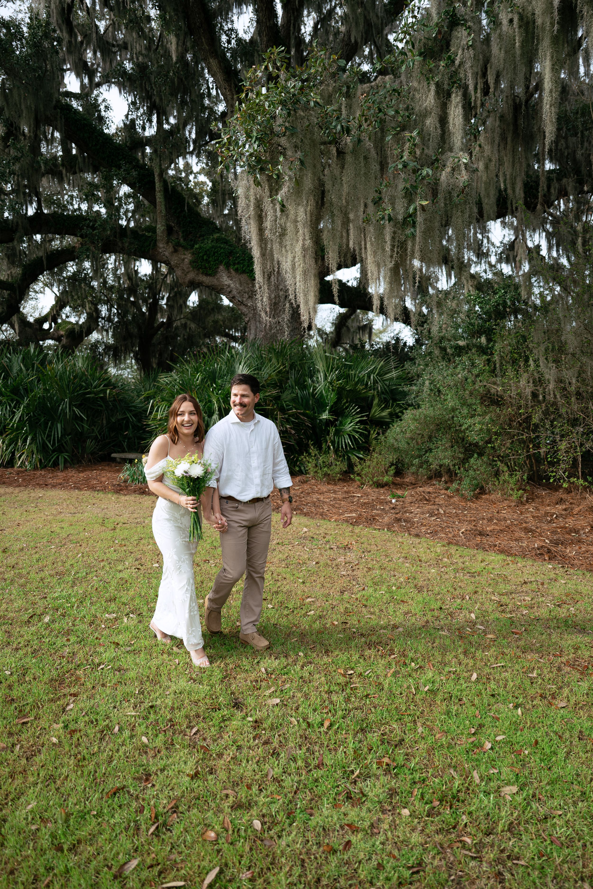 A couple in wedding attire poses on a grassy hill with Spanish moss-draped trees in the background at a Southern wedding.