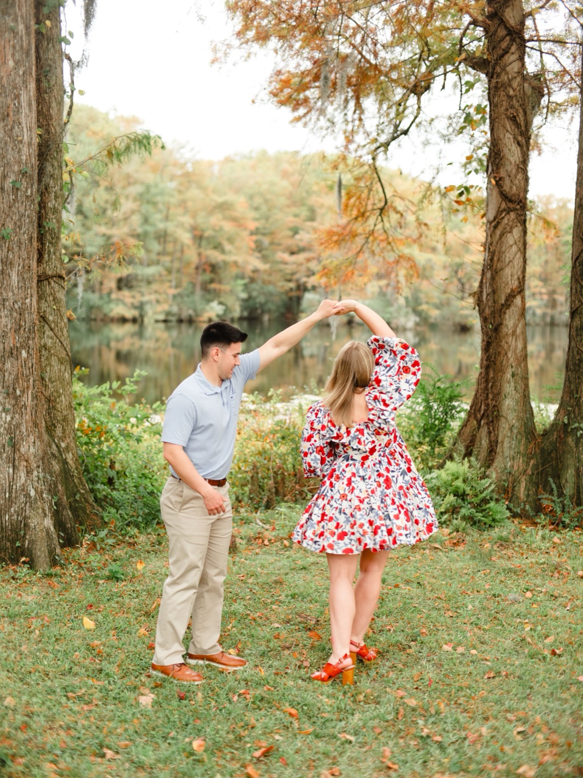 A couple dances together in a romantic autumn setting by a lake with orange and yellow fall foliage in the background.