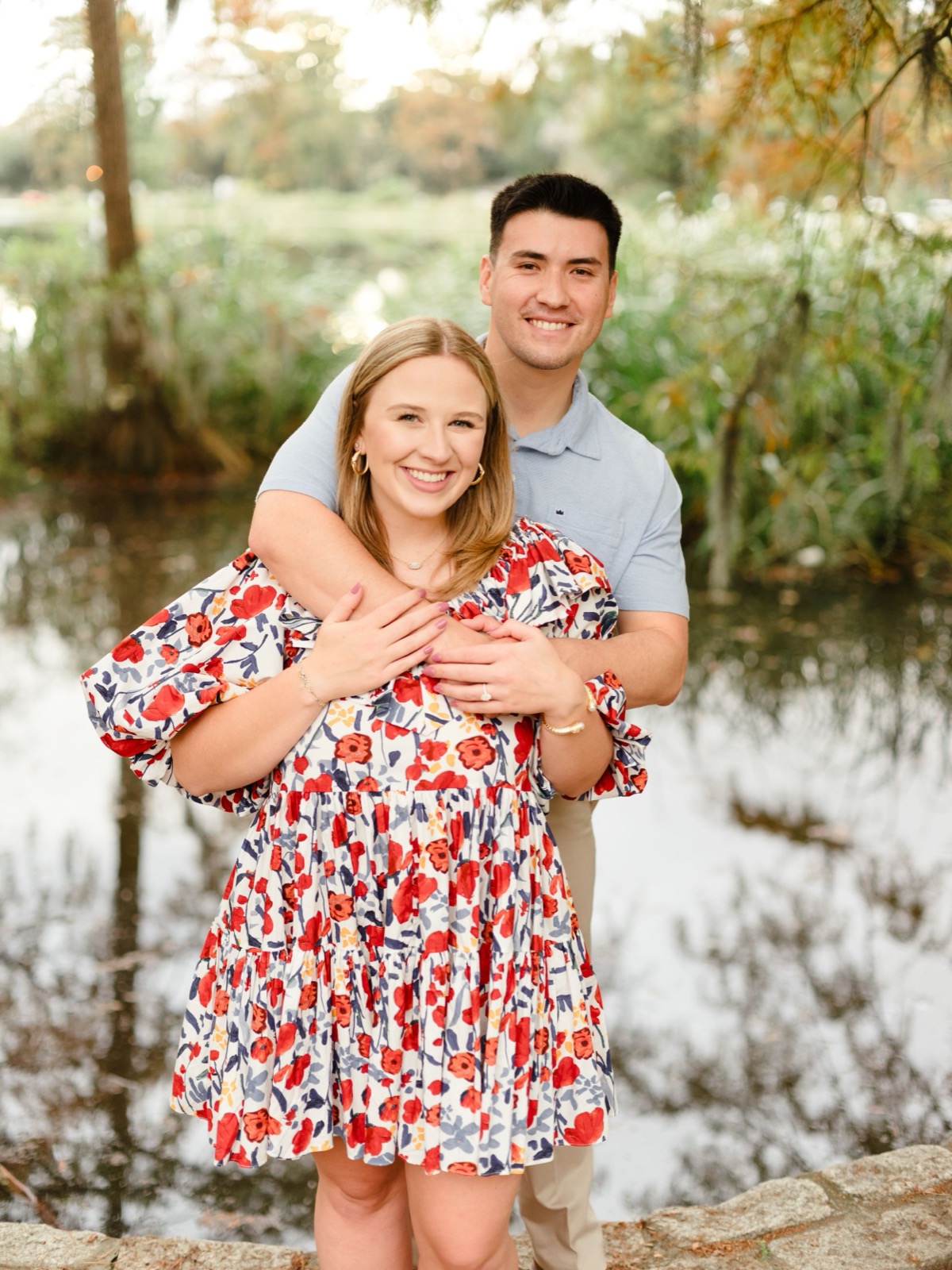 A couple embraces outdoors by a pond, with the woman wearing a red and white floral dress in a natural setting.