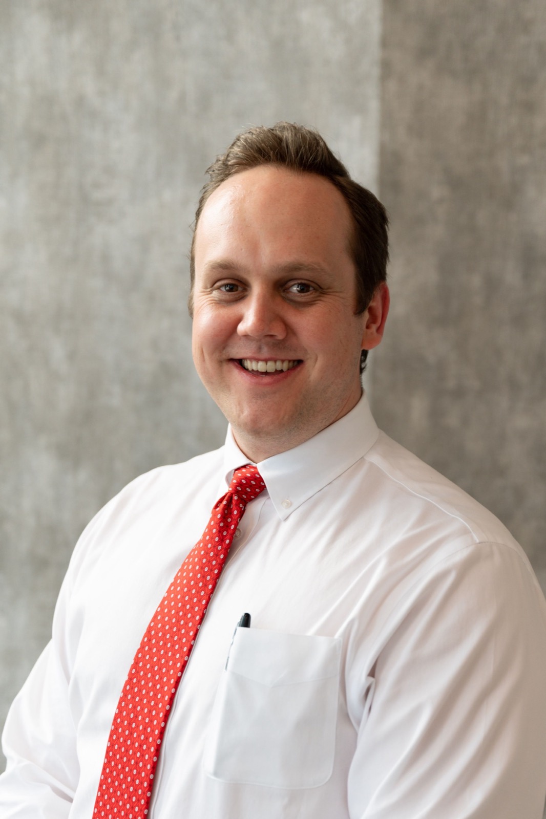 Professional portrait of a person in a white shirt and red tie against a gray background.