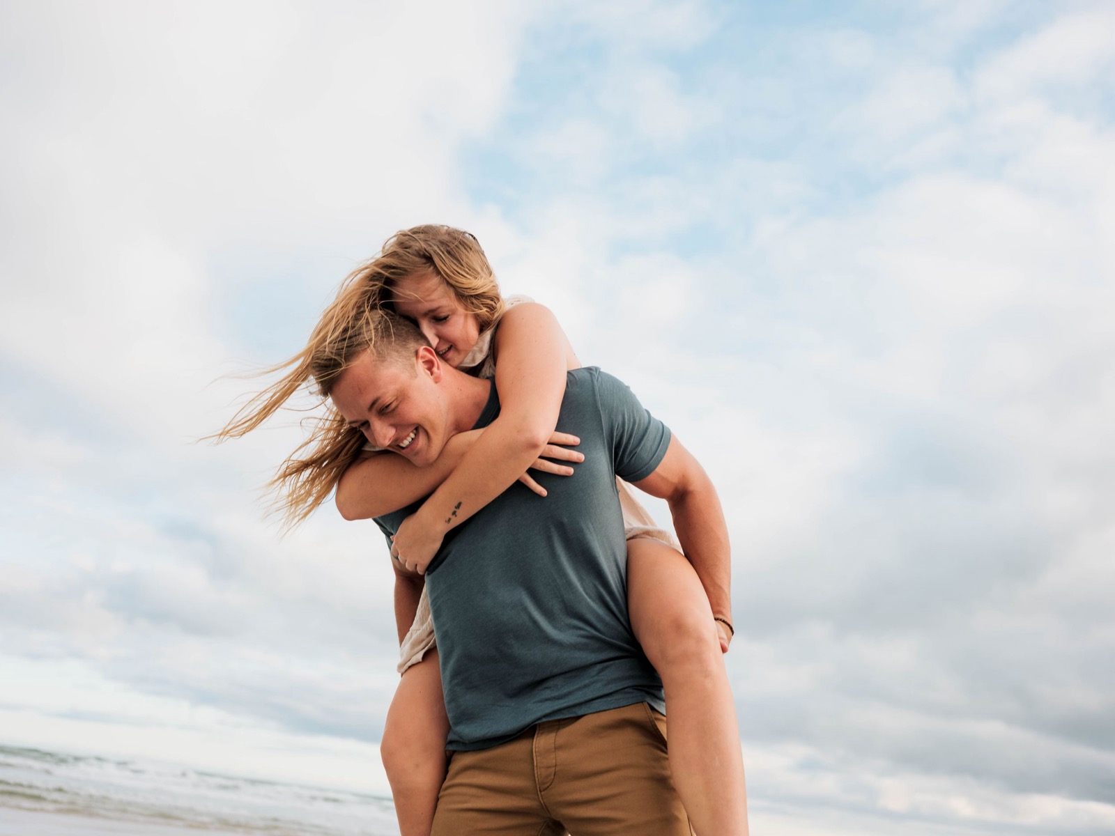 Playful couple enjoying a piggyback ride on cloudy beach.