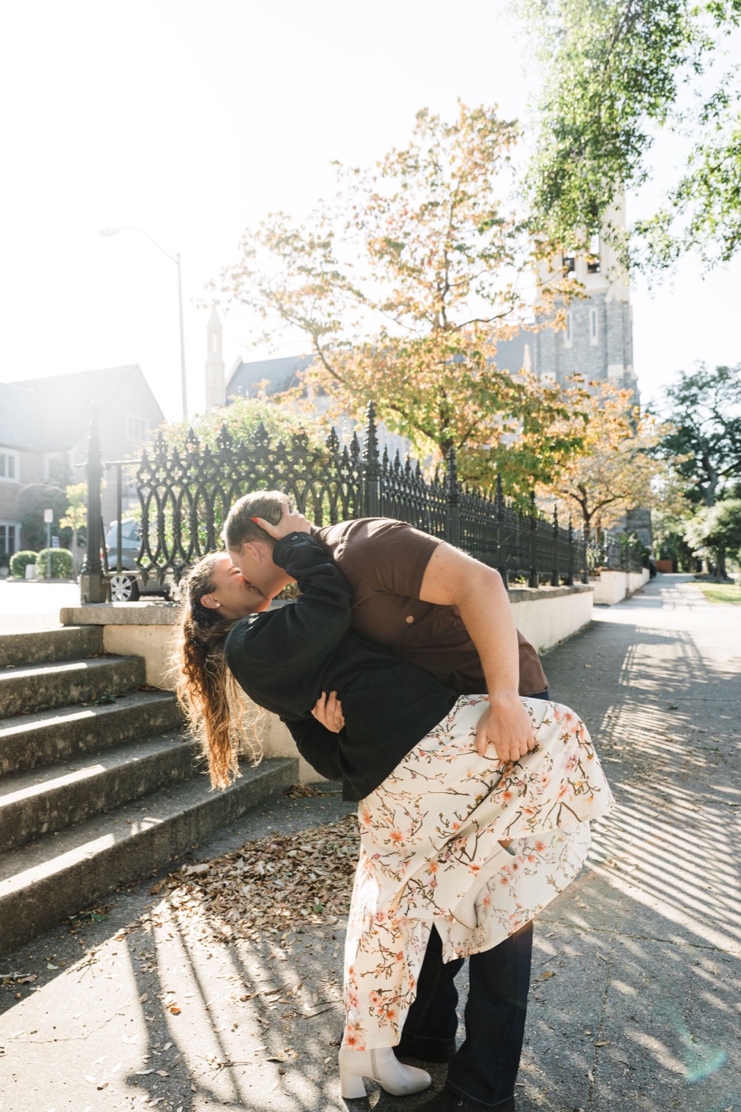 Romantic dip kiss between couple on sunlit steps with ornate iron fence and autumn leaves.