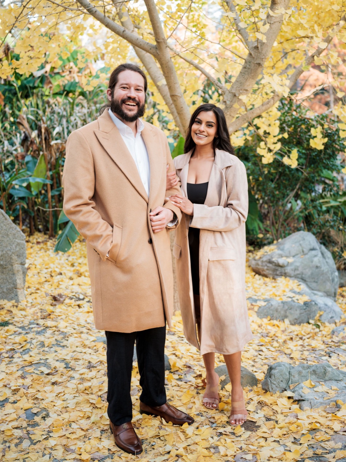 Couple in matching camel coats posing among autumn leaves in a garden setting.