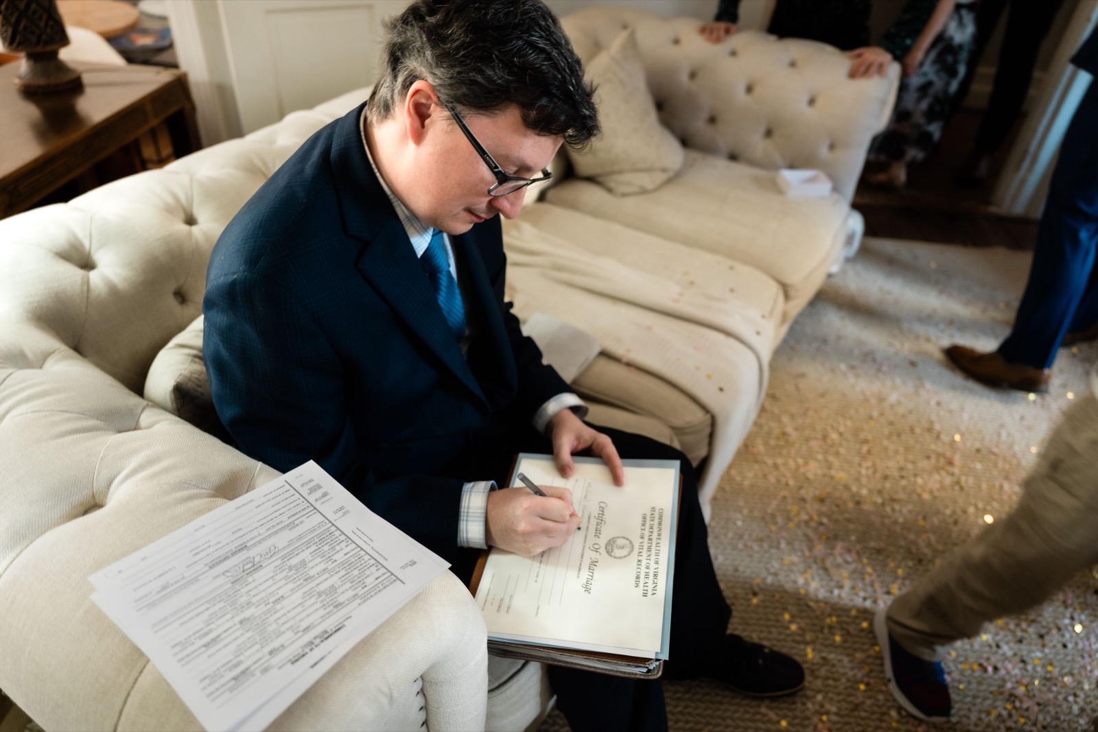 Someone in a navy suit sits on a beige couch while writing in a notebook during a quiet moment.