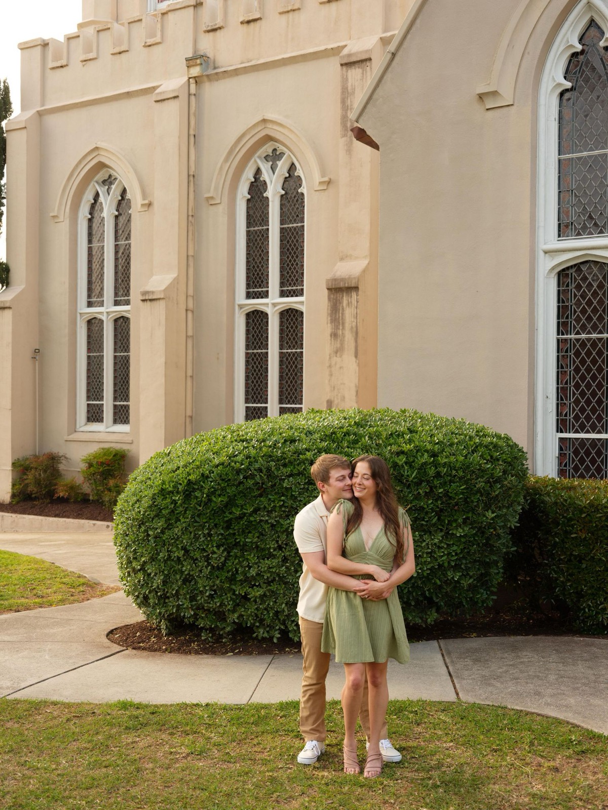 Romantic couple portrait in front of Gothic revival church with arched windows and manicured shrubs.