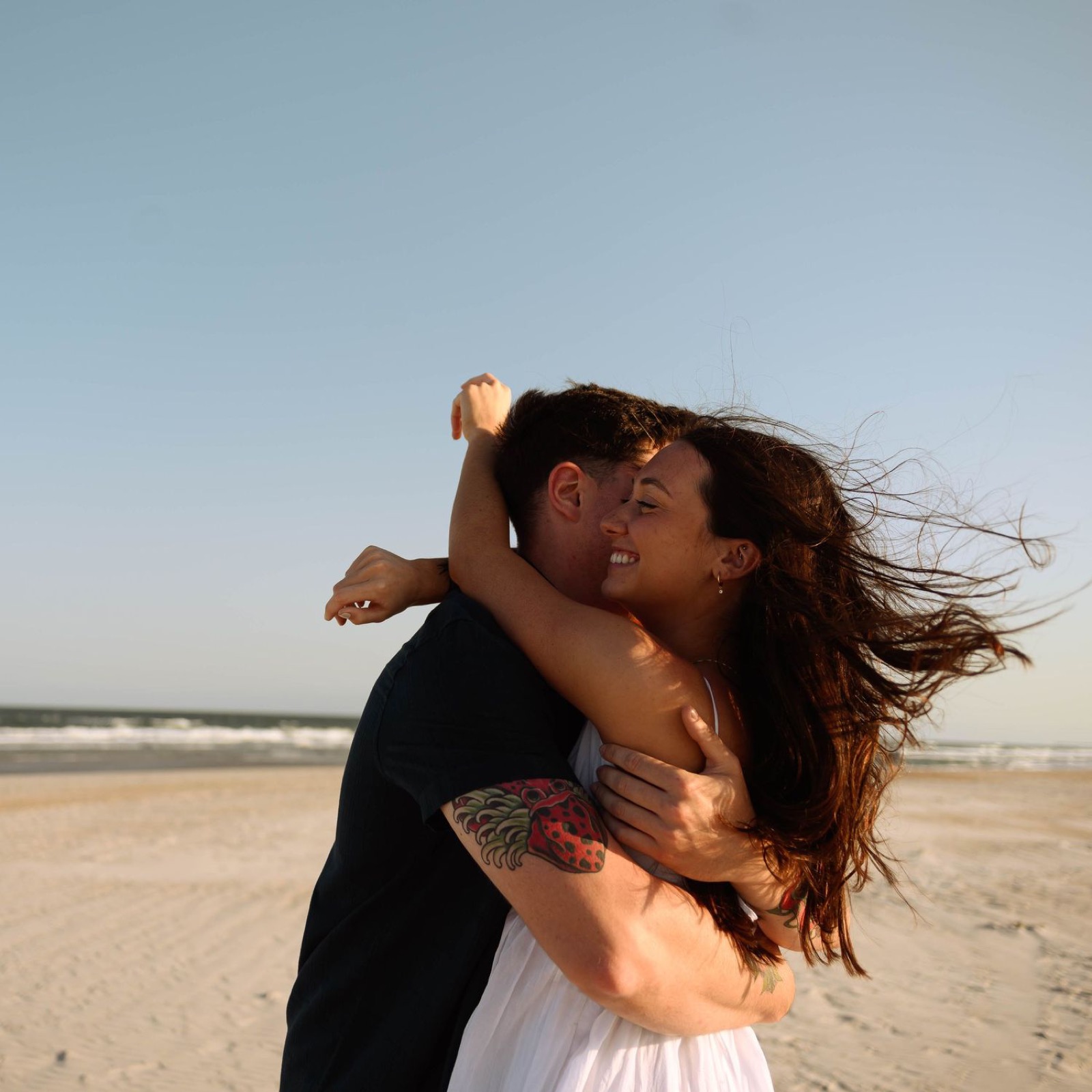 Playful beach photoshoot with couple dancing in wind at sunset.