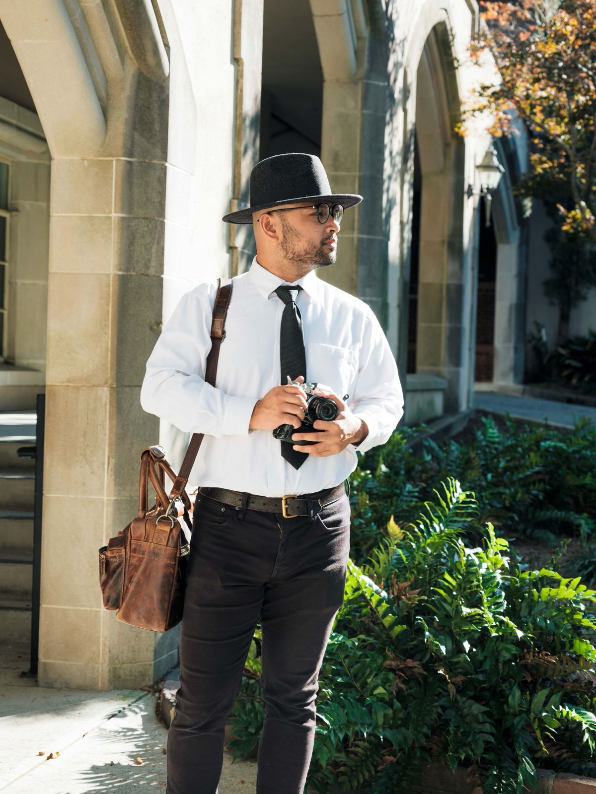 A well-dressed photographer in a fedora and white shirt holds a vintage camera in an elegant archway.
