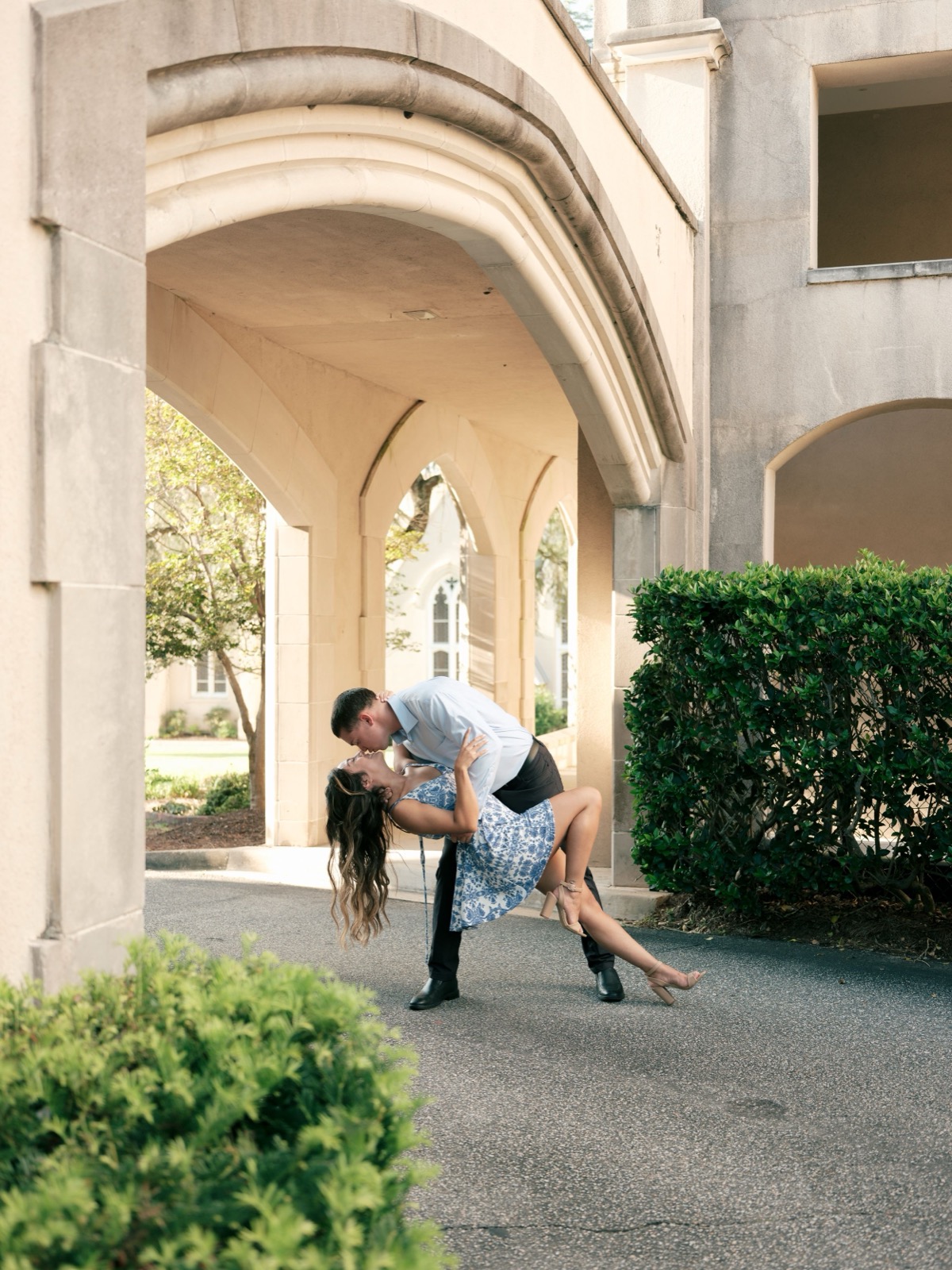 A romantic dance pose captured under arched colonnade with decorative hedges and architecture in the background.