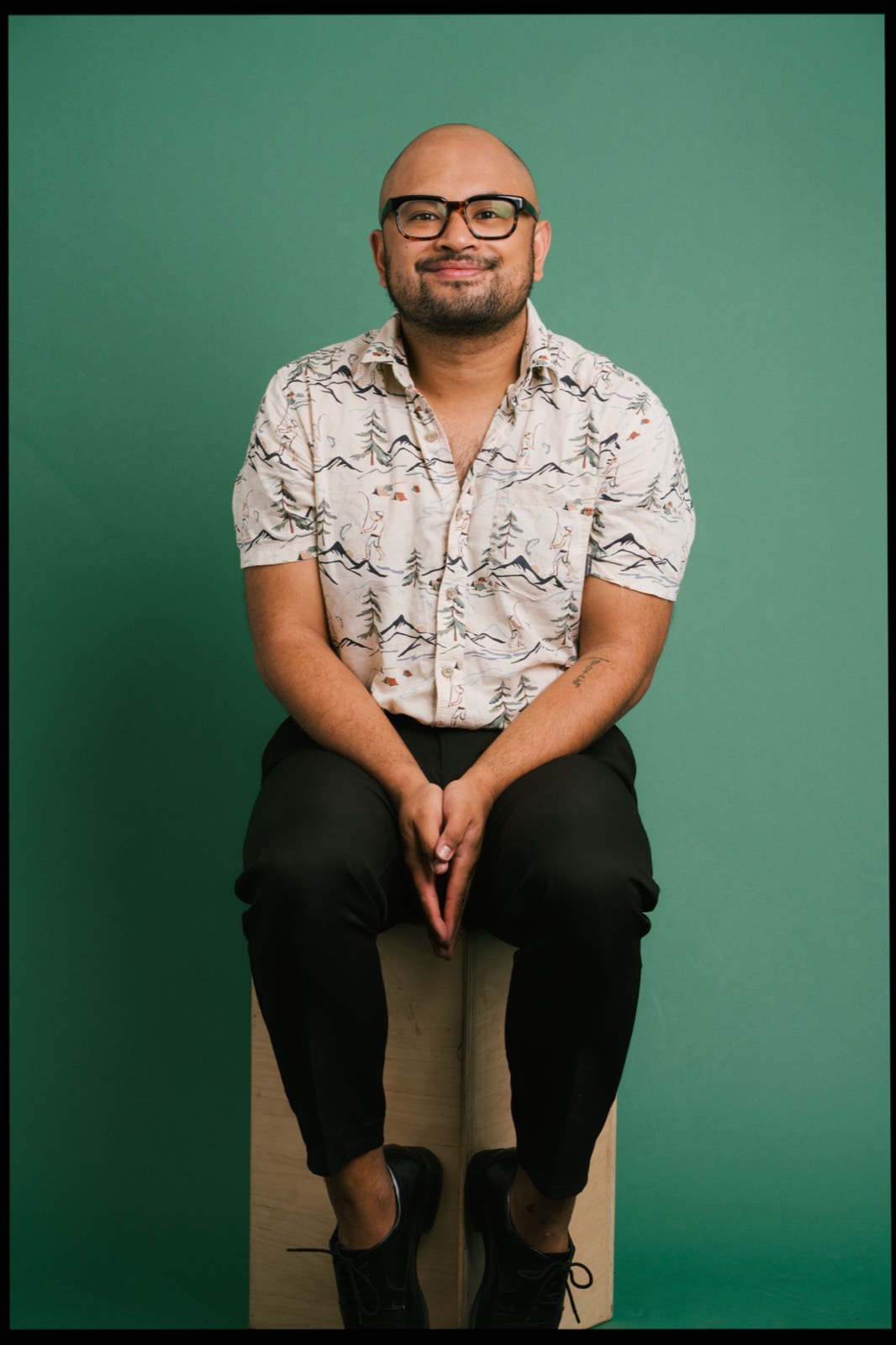 Portrait against a green backdrop of someone wearing glasses, a patterned short-sleeve shirt and black pants.