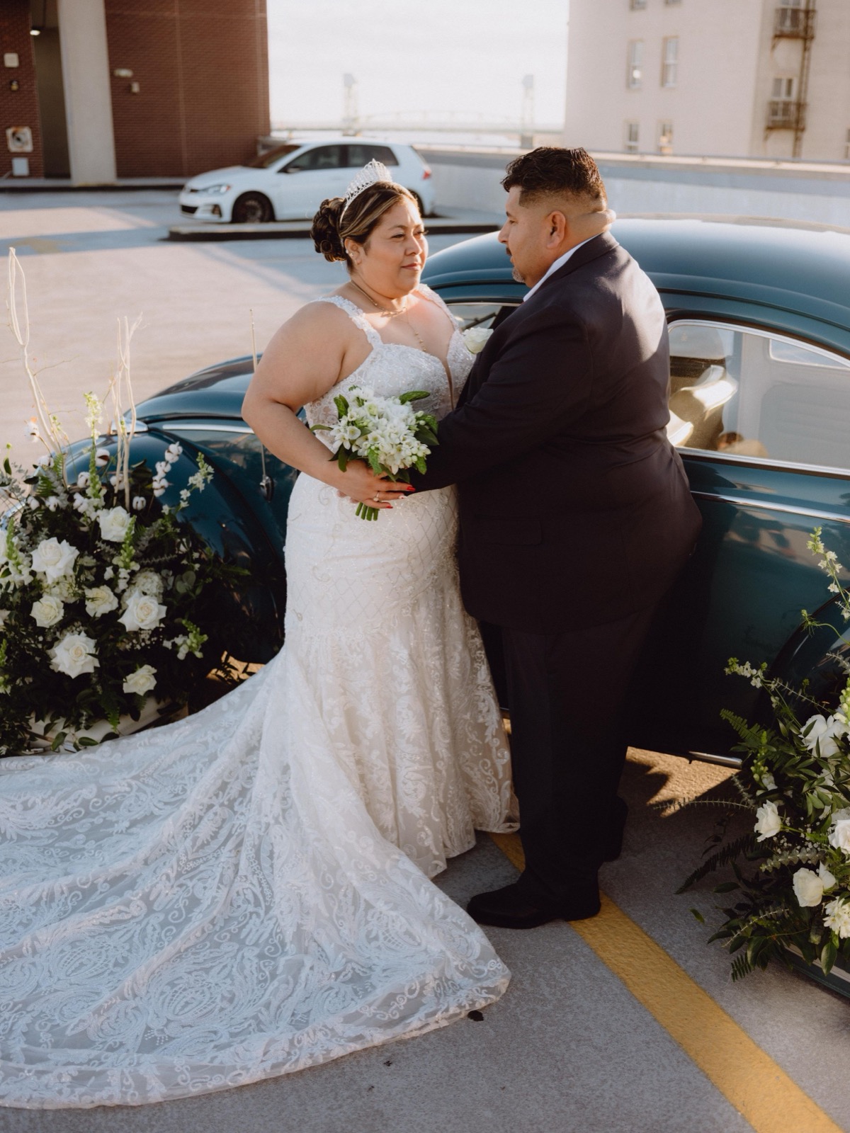 Wedding couple posing by decorated vintage car with white floral arrangements.