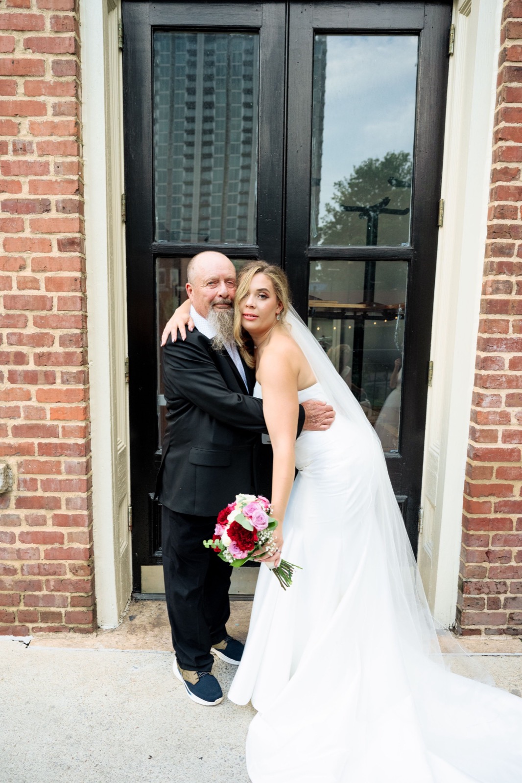 Wedding couple poses together in a doorway of a brick building with the bride holding a pink bouquet.