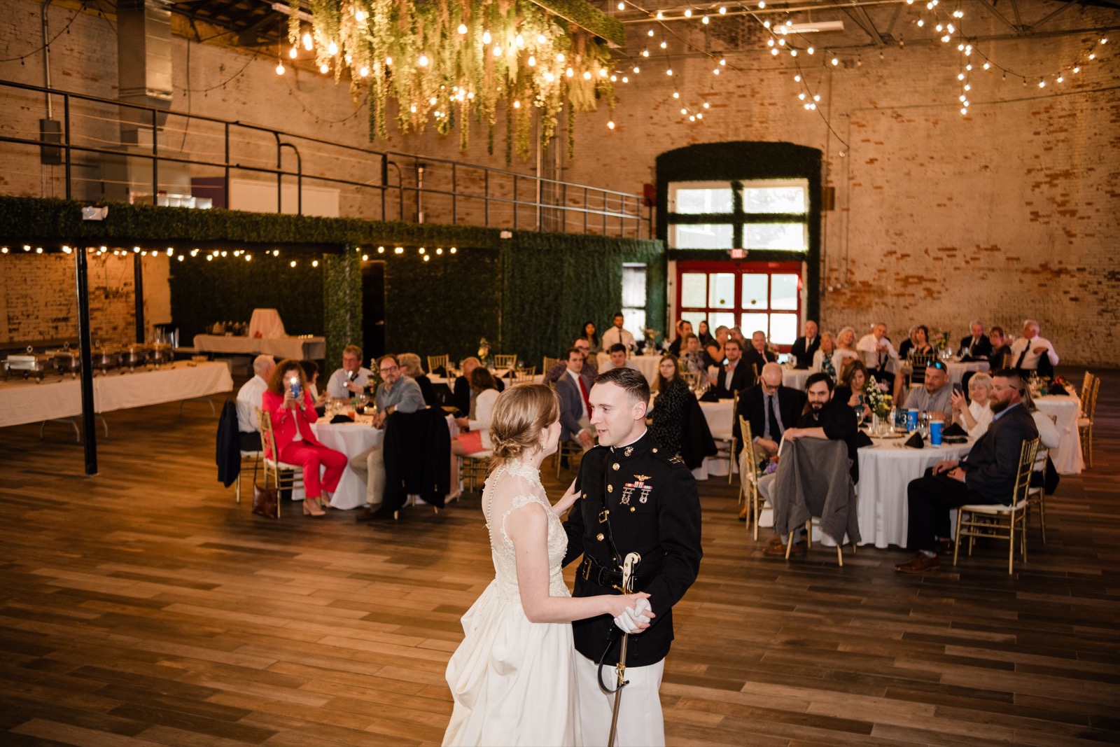 Indoor wedding reception scene with guests seated at round tables under string lights and chandeliers in a rustic brick venue.