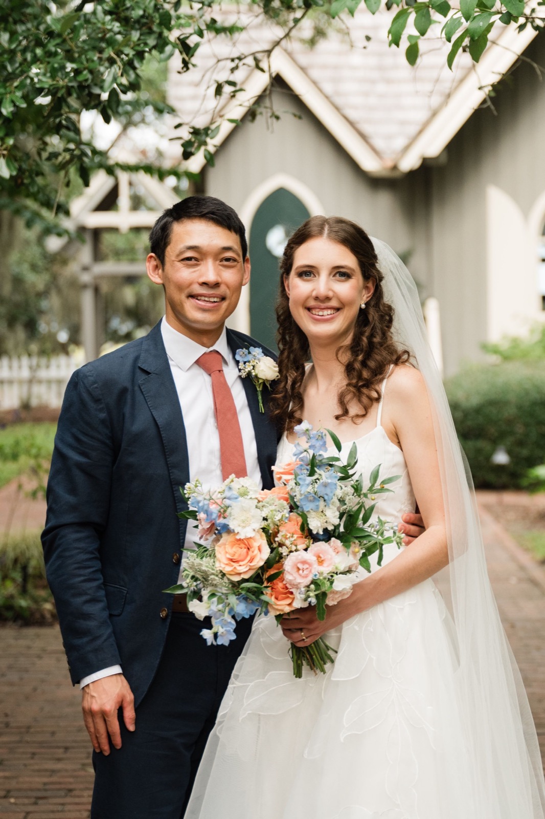 A newlywed couple poses outdoors at their wedding with a beautiful peach and blue floral bouquet.