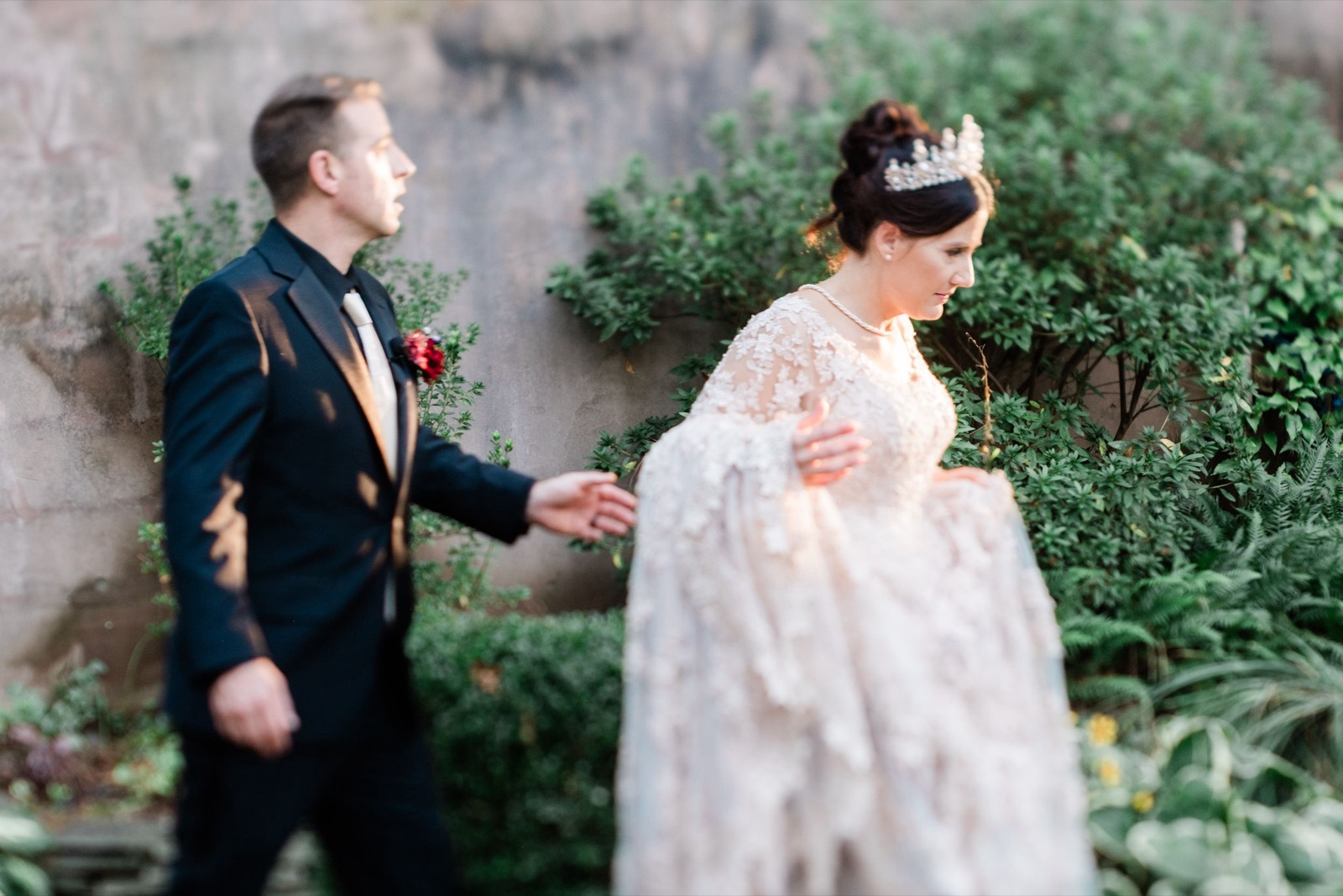 A dreamy first look wedding moment between bride in lace gown and groom in black suit amid garden greenery.