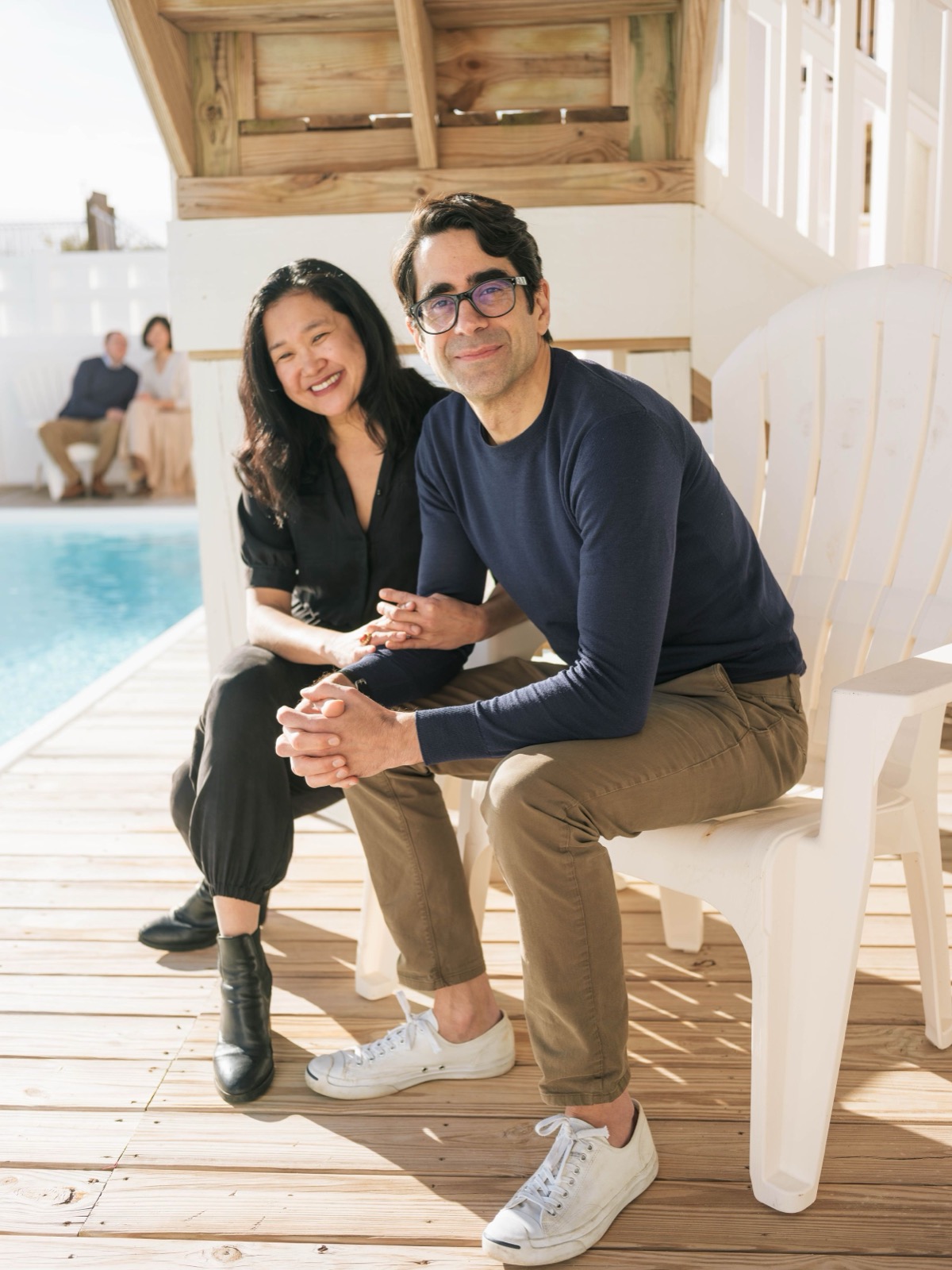 A couple sitting on white outdoor furniture by a swimming pool.