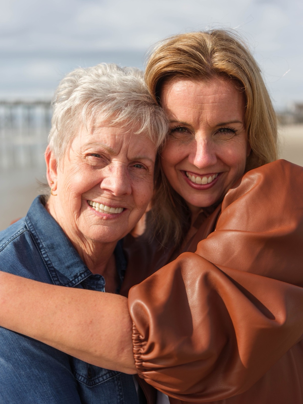 Two generations share a warm embrace on a beach with a pier in the background.