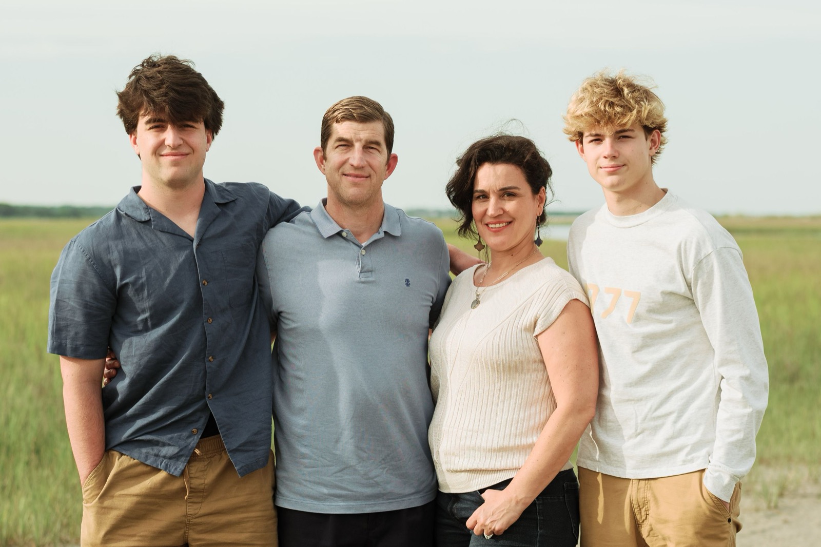A family of four poses together in casual attire against a natural outdoor background.
