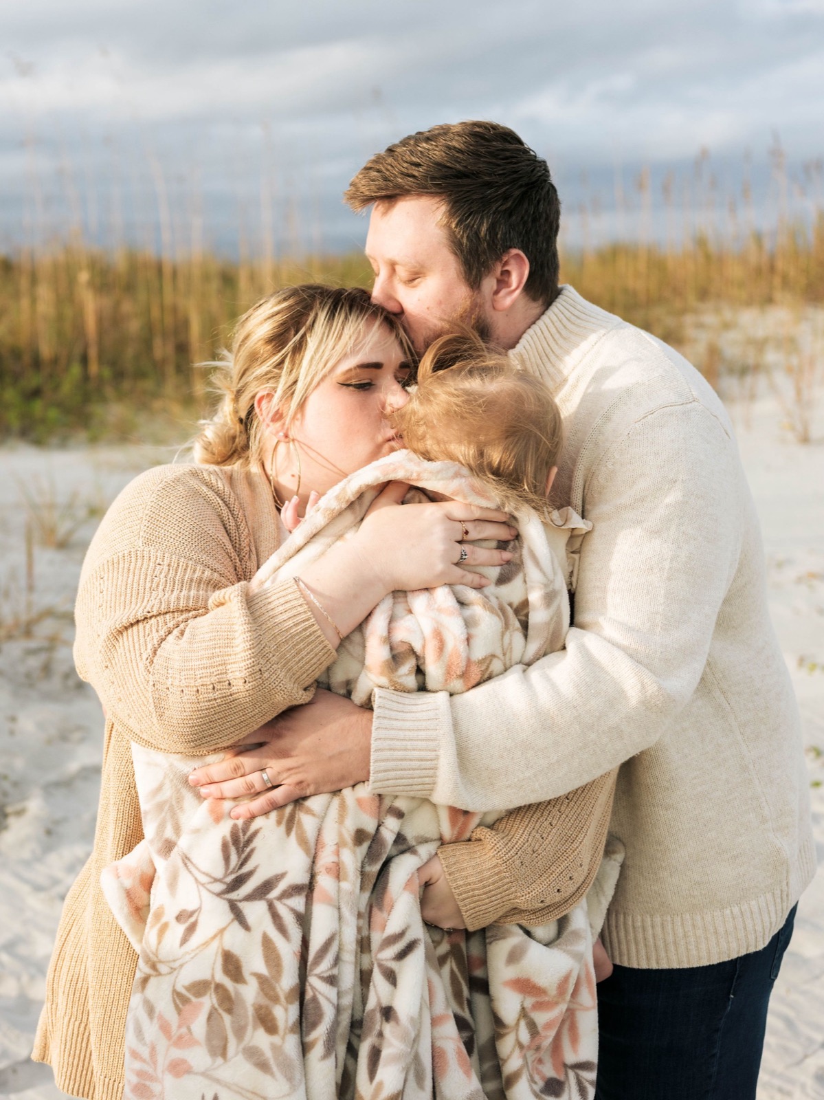 A couple in cozy sweaters sharing an intimate moment while holding a blanket at the beach.