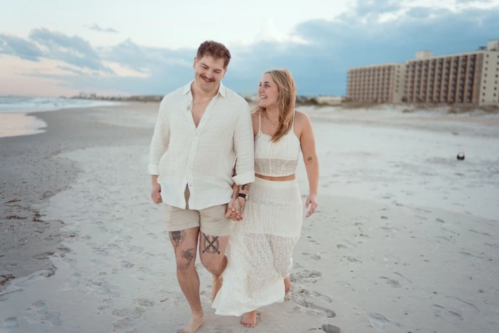 Couple in white outfits walking together on a sandy beach at sunset with resort buildings in the background.