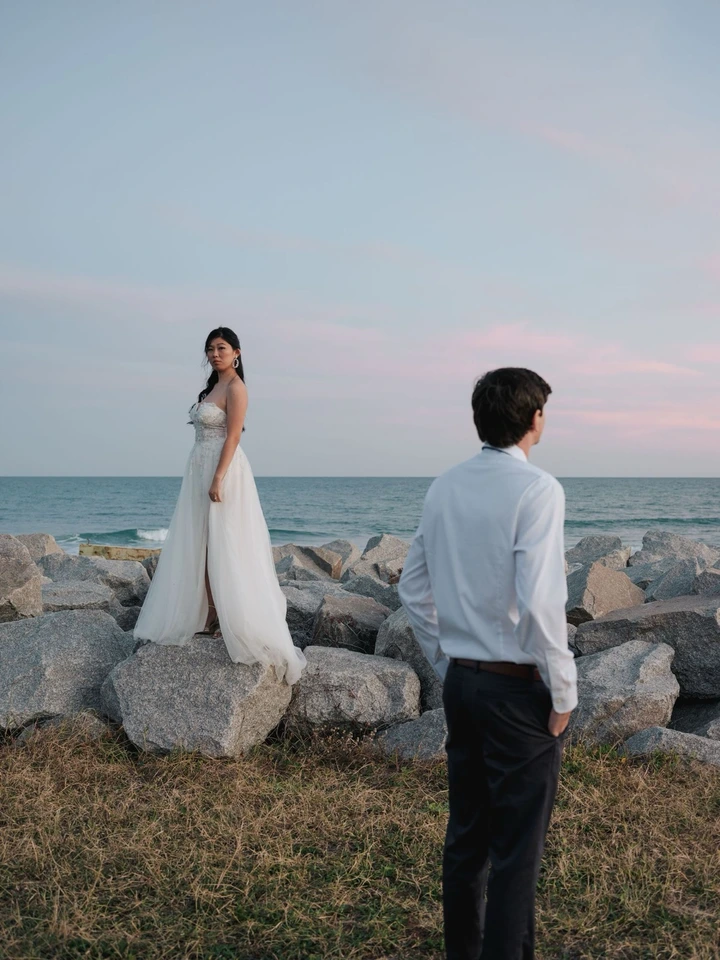 A couple in wedding attire poses on rocky shoreline during sunset with ocean in background.